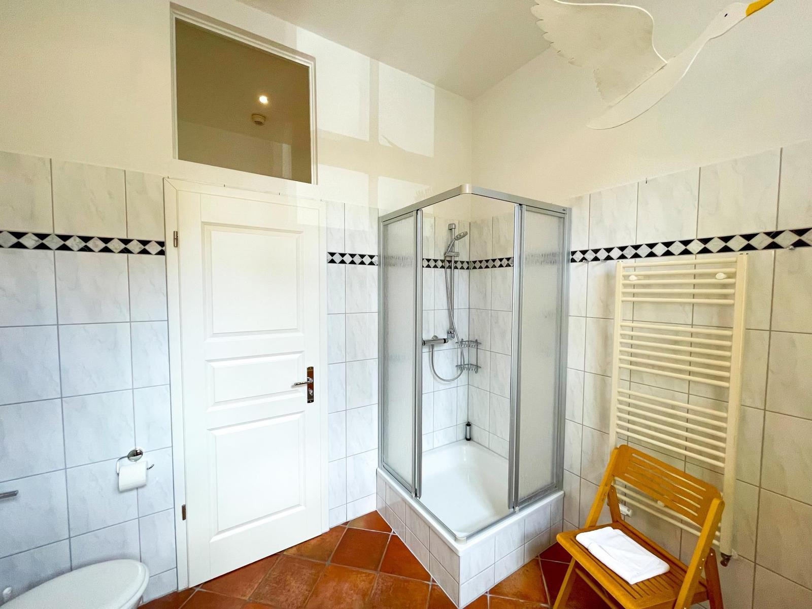 Badezimmer mit Dusche, Toilette und Heizkörper. Holzstuhl und Vogel an der Decke.
