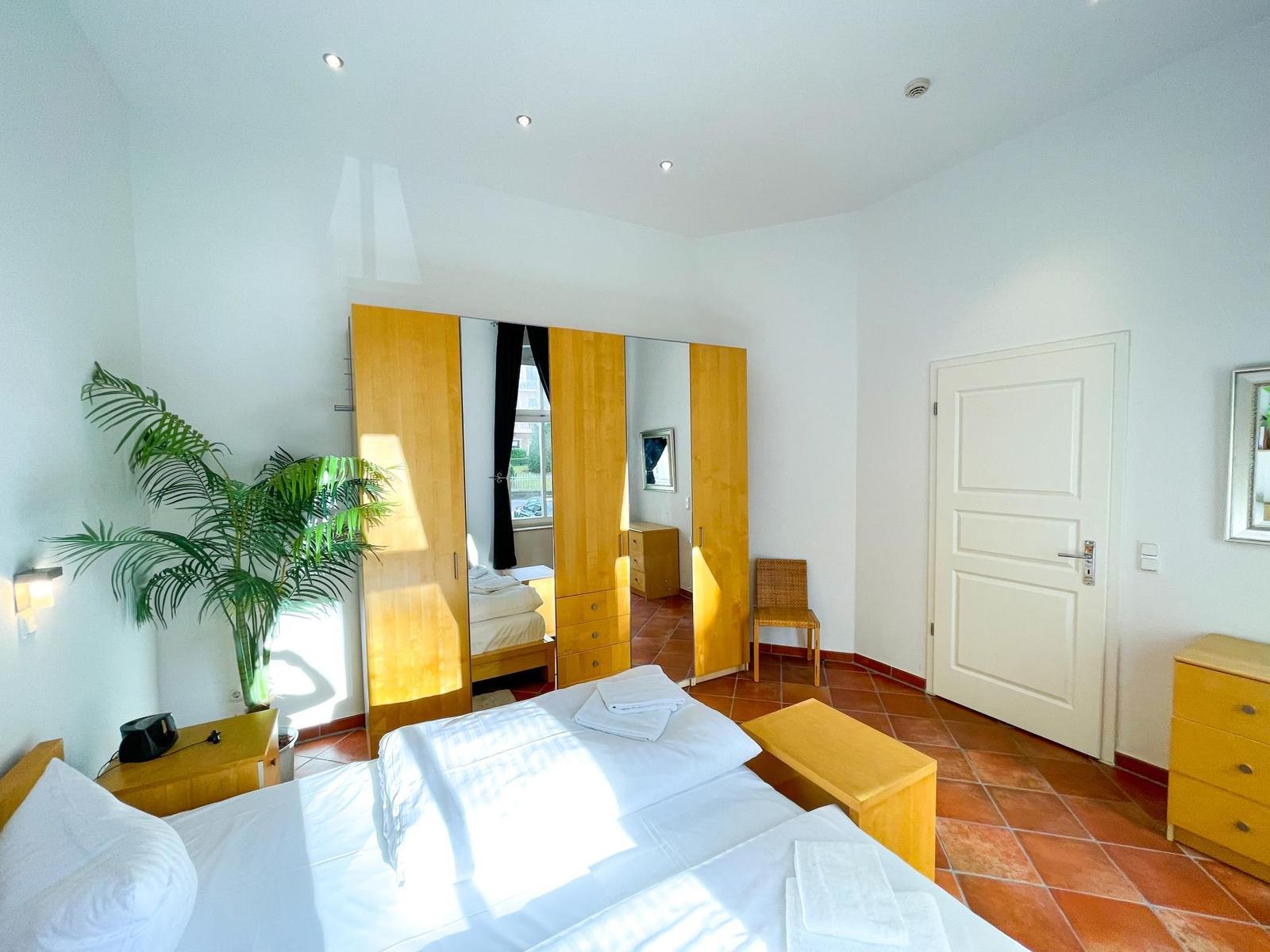 Schlafzimmer mit Doppelbett, Spiegelschrank, Pflanze und Terrakottboden.