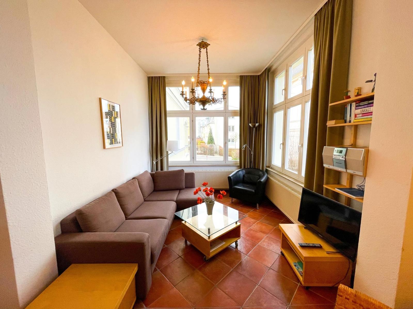 Wohnzimmer mit Sofa, Glas-Tisch und Fernseher. Große Fenster und Kronleuchter.