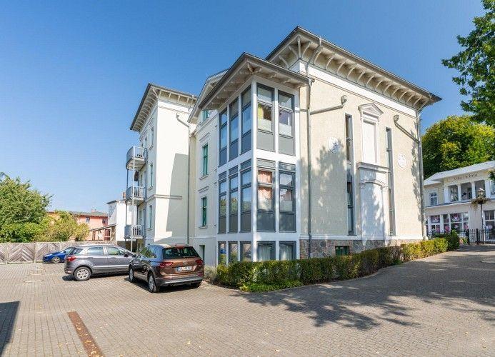Haus mit mehreren Etagen, großen Fenstern und gepflastertem Hof mit parkenden Autos.