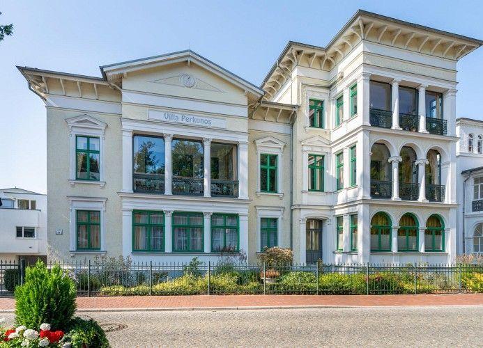 Villa Perkunos mit grünen Fensterrahmen und Balkonen, umgeben von Garten und Zaun.