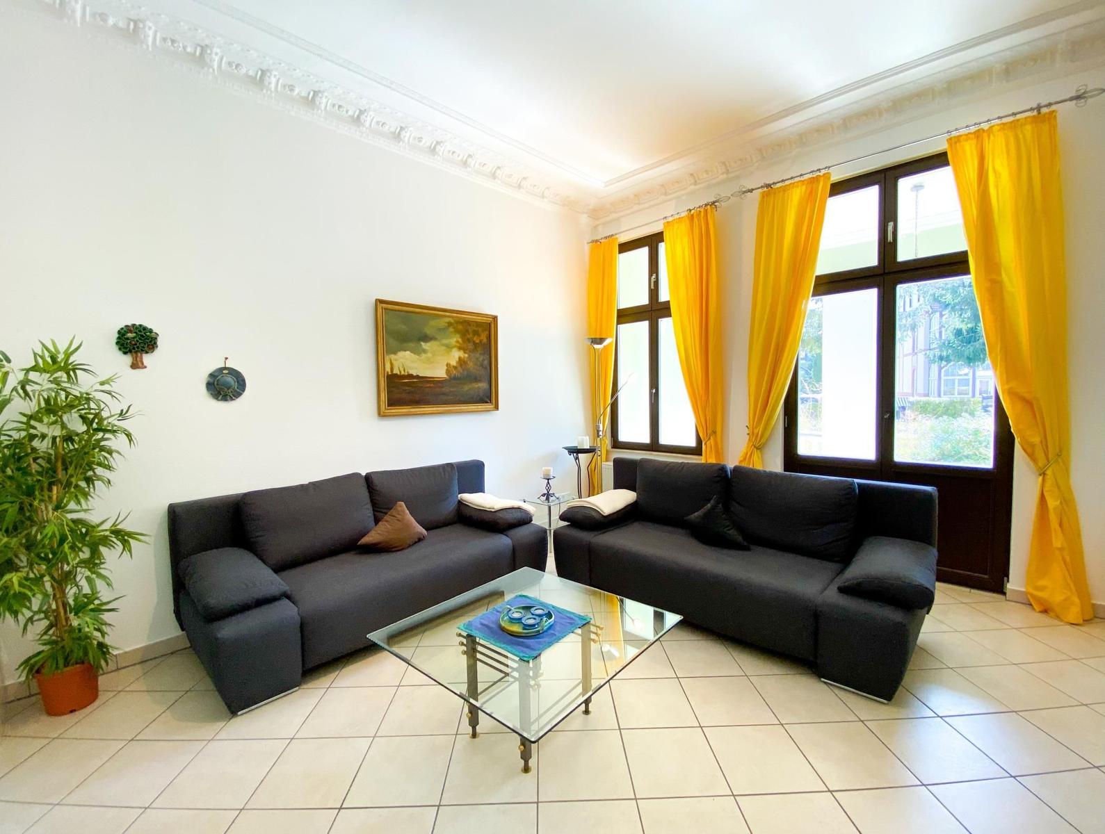 Zwei schwarze Sofas um einen Glas-Tisch. Gelbe Vorhänge und ein Bild an der Wand.