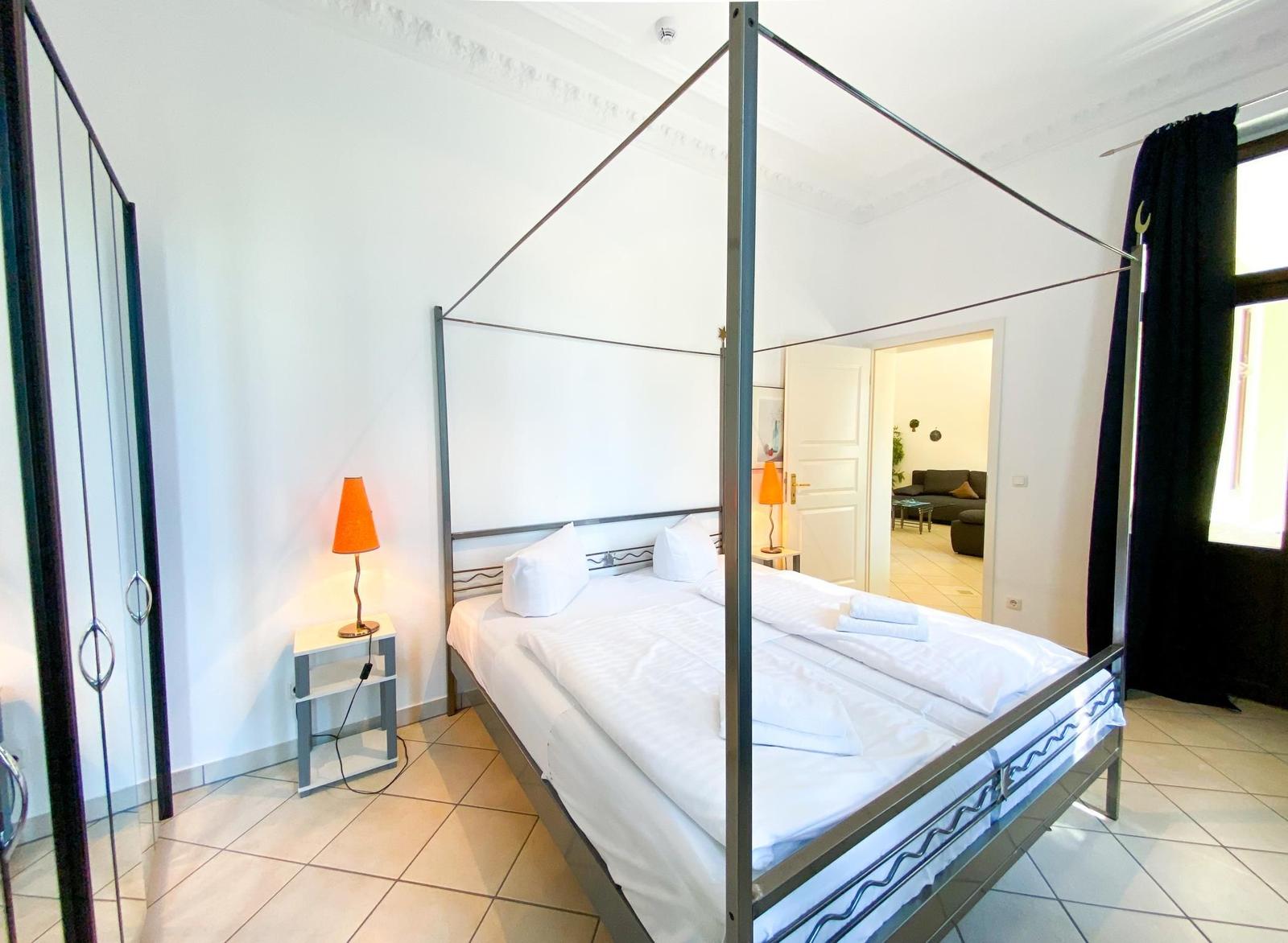 Schlafzimmer mit Doppelbett, Nachttisch und Lampe. Tür führt in Wohnbereich.