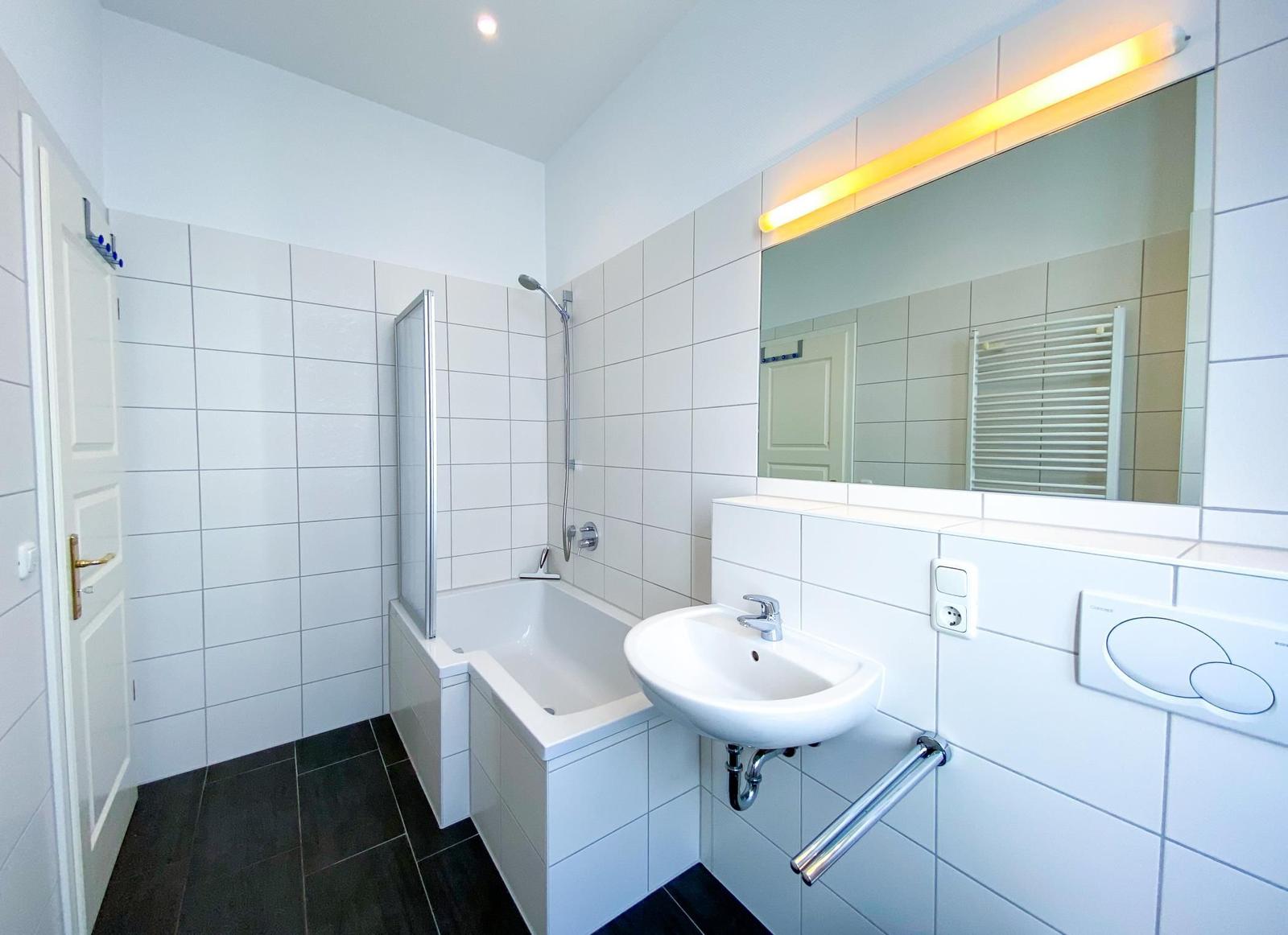 Badezimmer mit Dusche, Waschbecken und Spiegel. Fliesenwand und dunkler Boden.