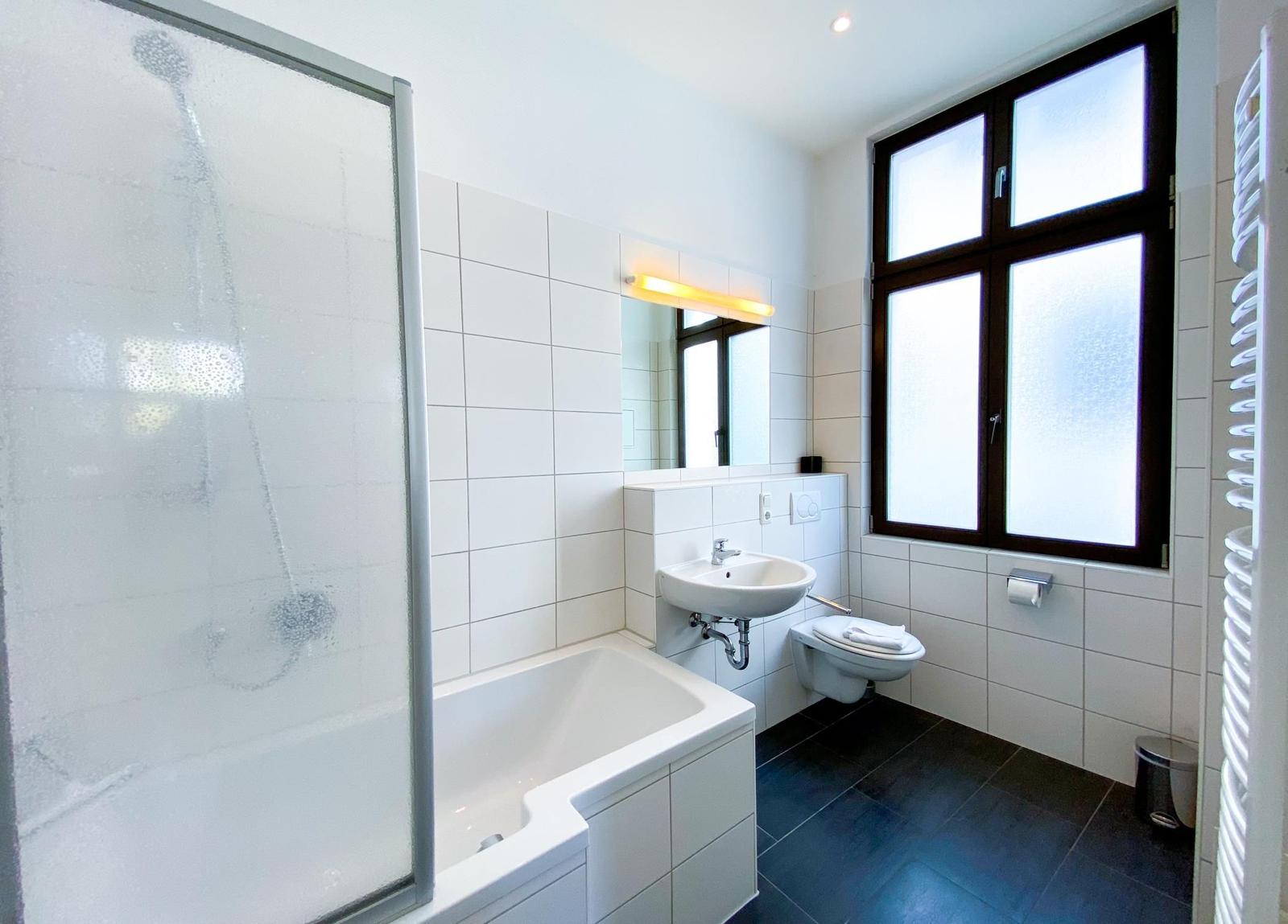 Badezimmer mit Badewanne, Dusche, Waschbecken und Toilette. Große Fenster mit Milchglas.