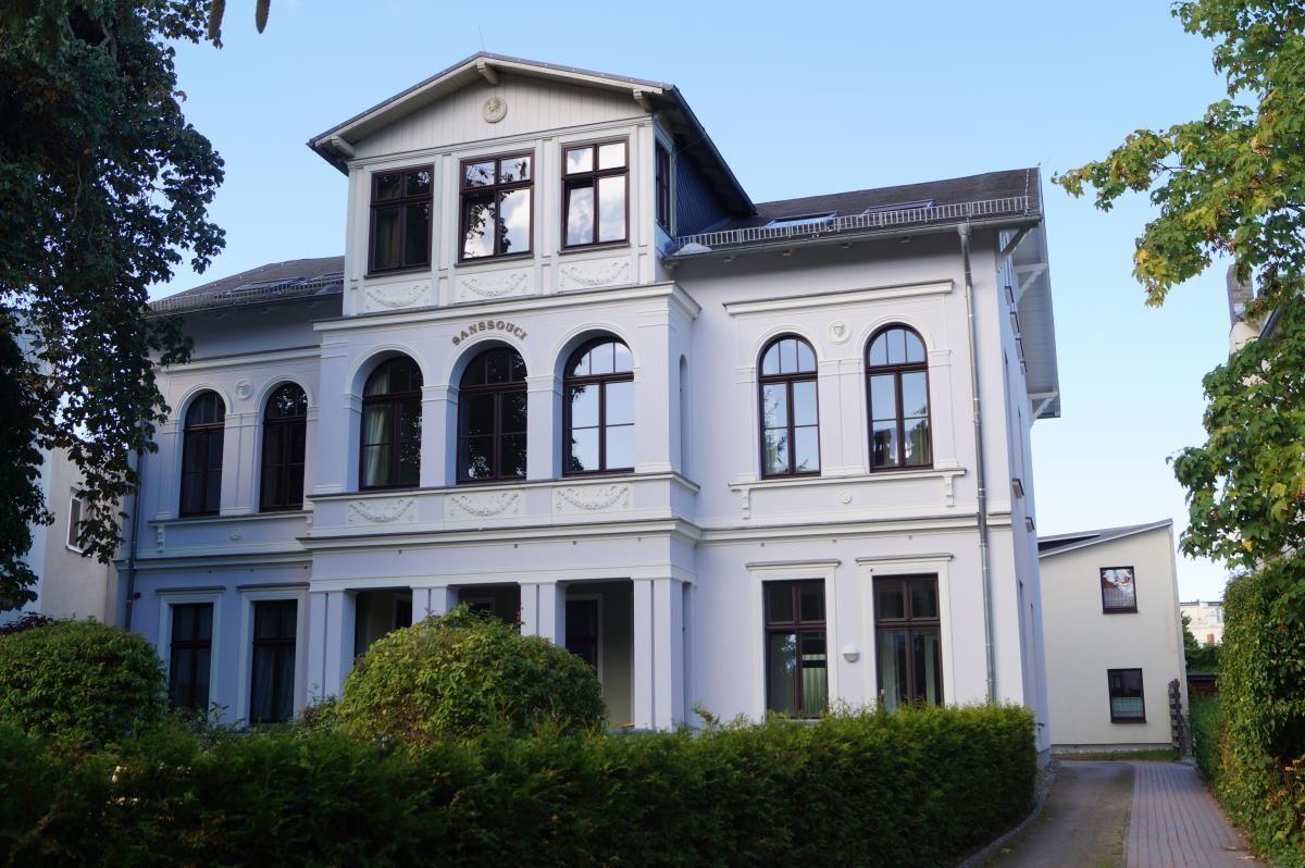Ein weißes, dreigeschossiges Haus mit Bogenfenstern und einem Balkon. Umgeben von grünen Sträuchern.