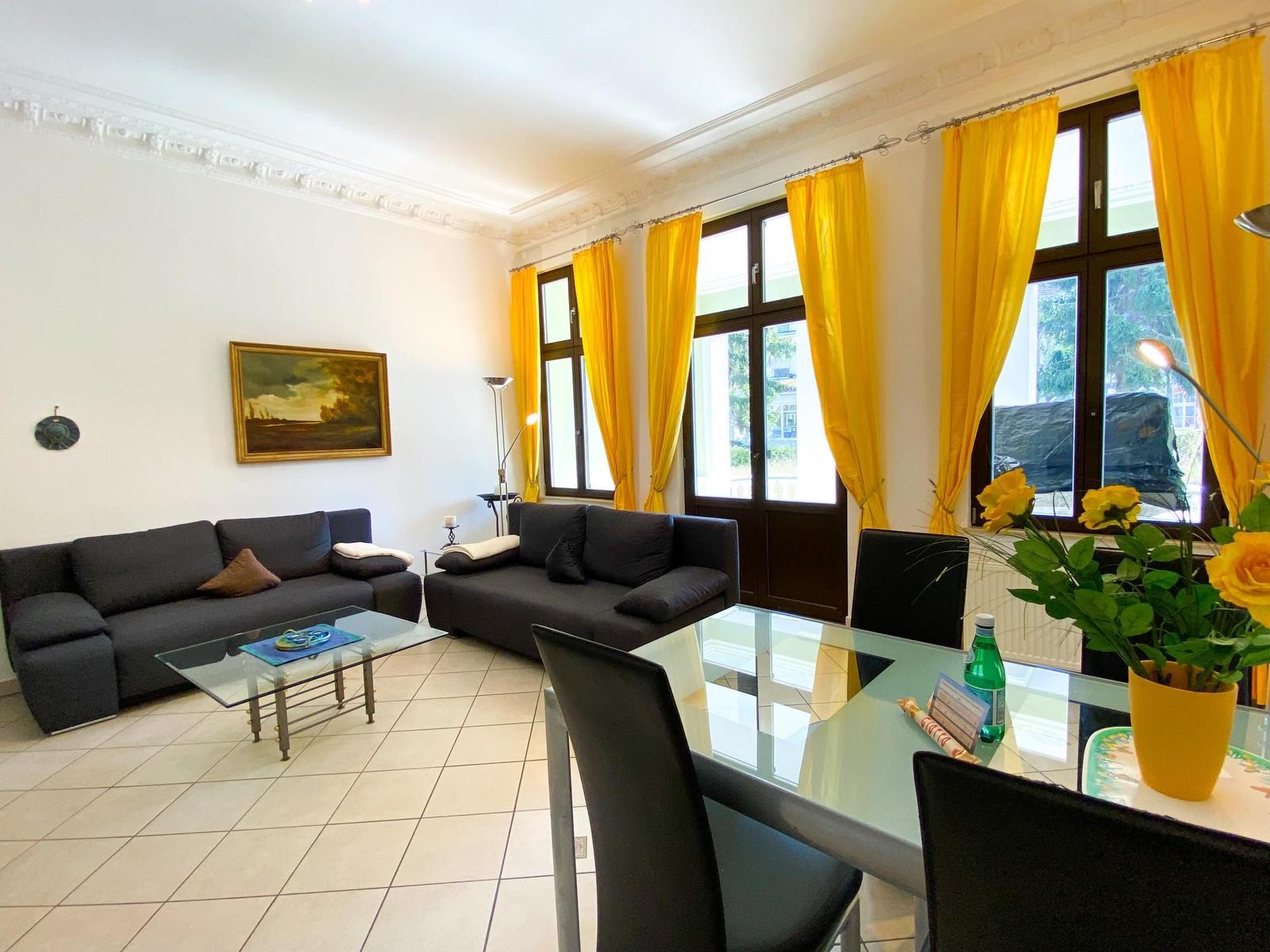 Wohn- und Essbereich mit zwei Sofas, Glas-Tisch und gelben Vorhängen.