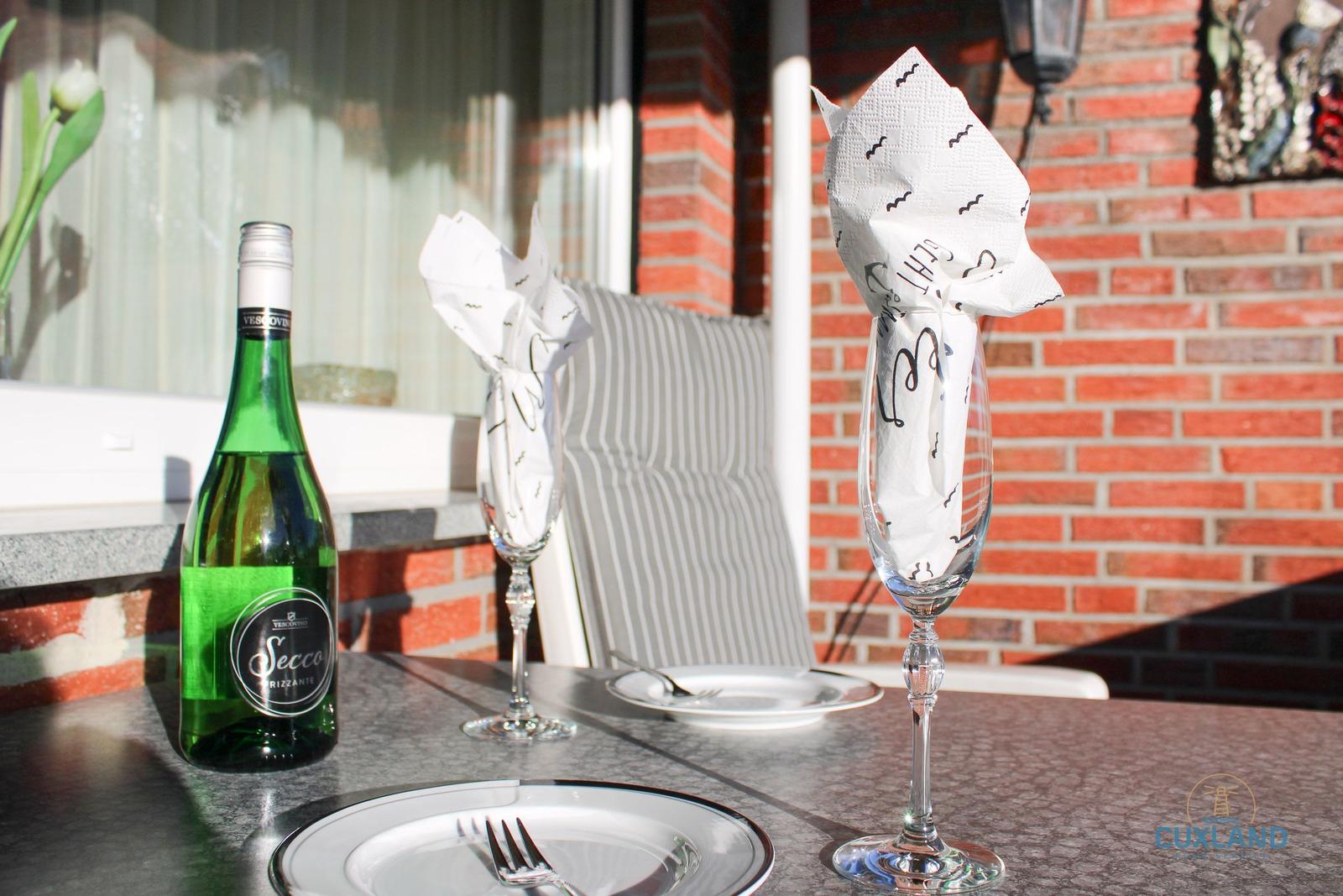 Tisch mit Flasche Secco, zwei Gläsern und Tellern auf Terrasse mit Backsteinwand.