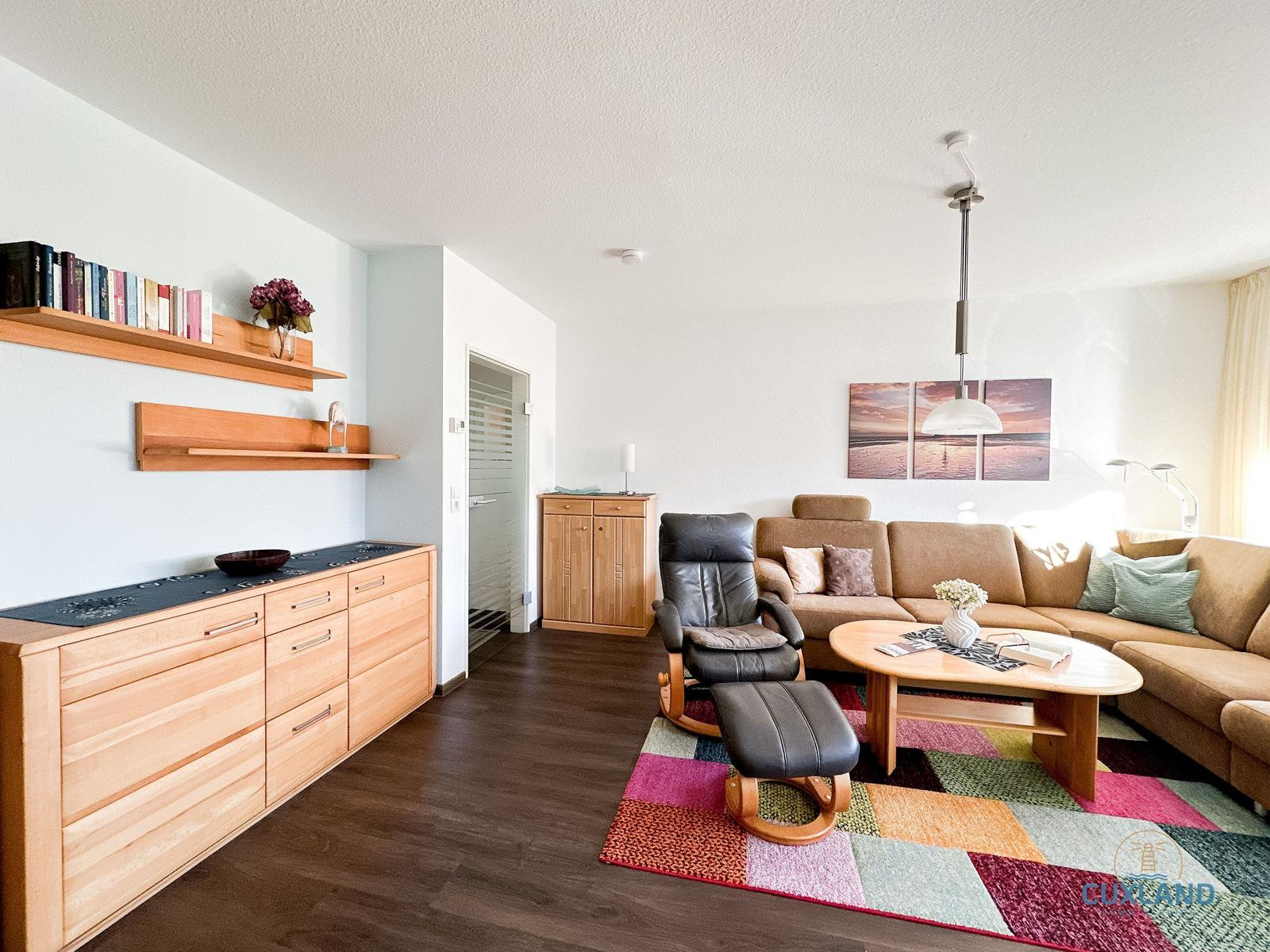 Wohnzimmer mit Sofa, Sessel, Tisch und Regal. Teppich und Wandbild sichtbar.