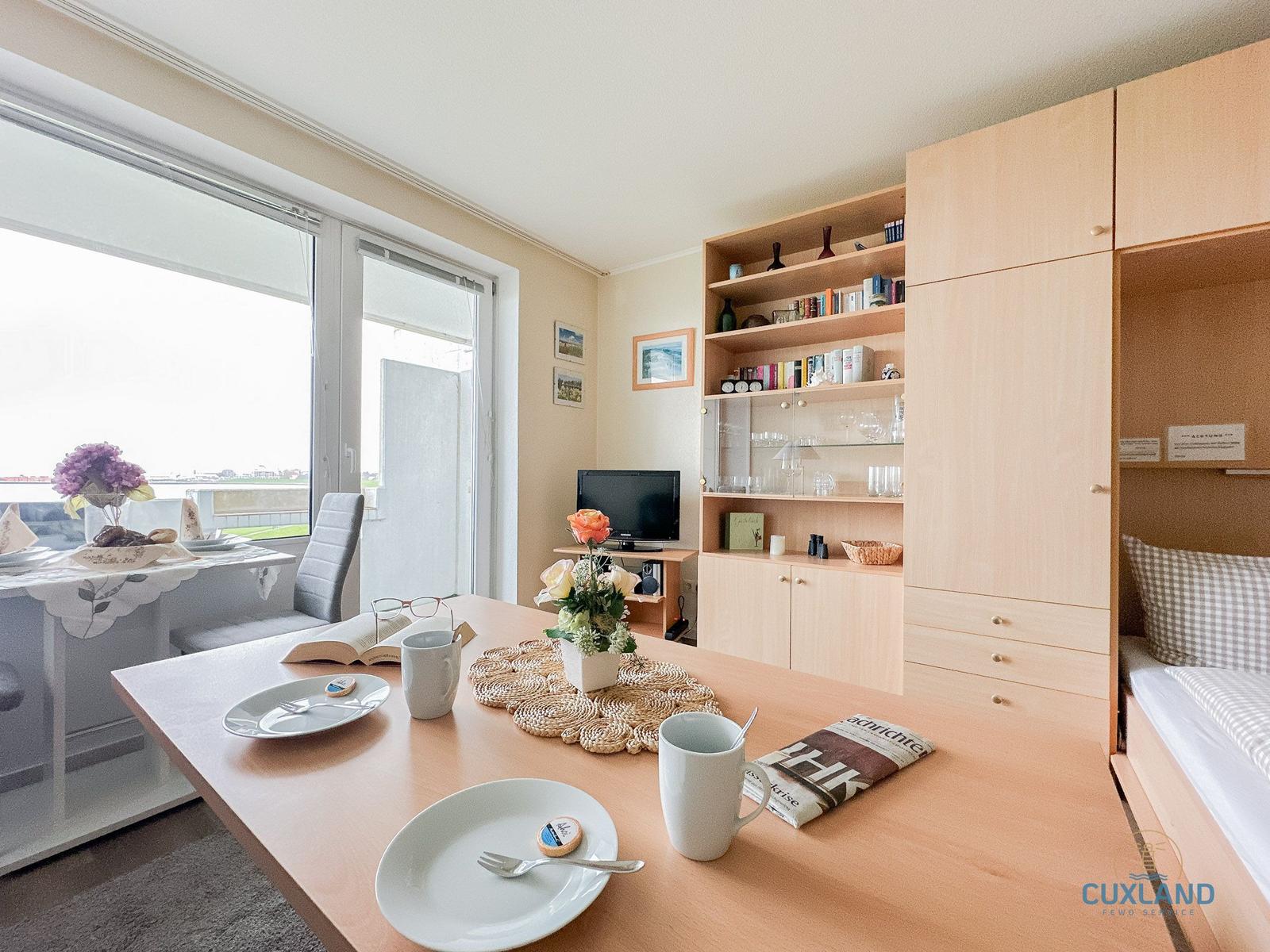 Essbereich mit Tisch, Stühlen und Blick auf Balkon. Hintergrund: Wohnbereich mit TV und Schrank.