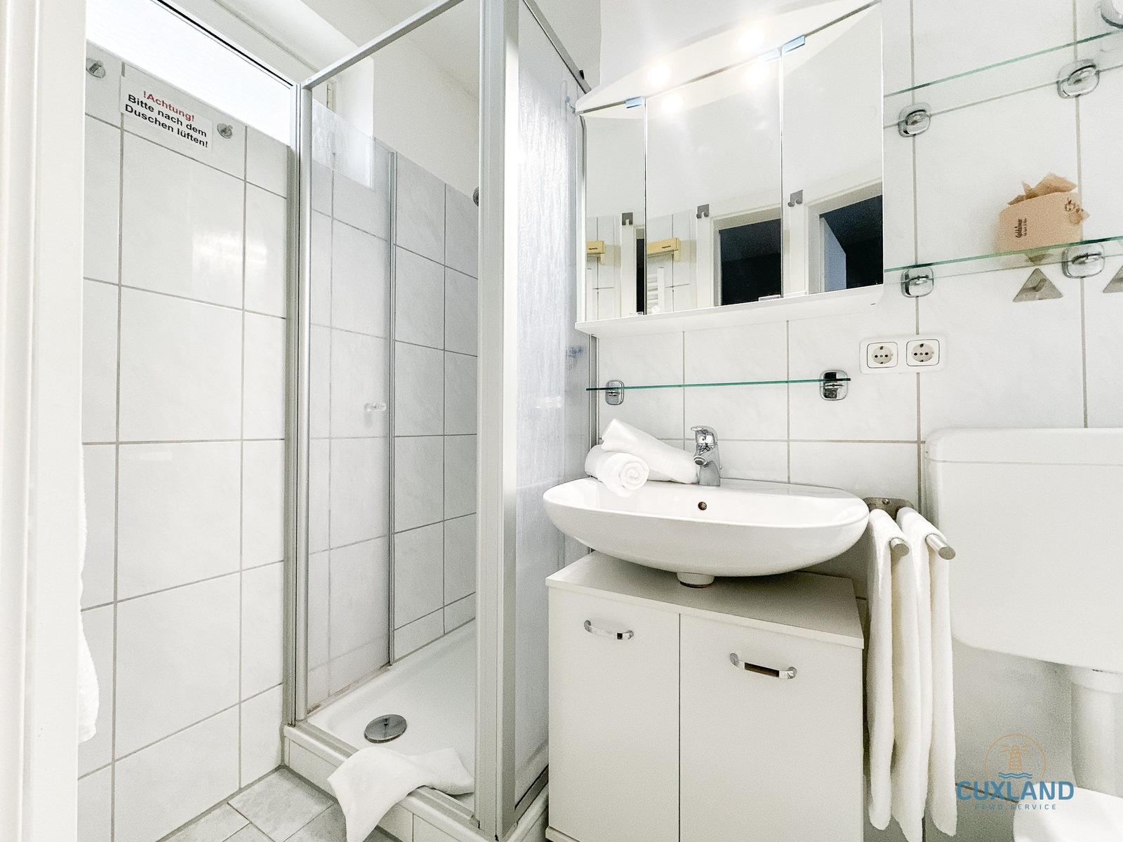 Badezimmer mit Dusche, Waschbecken und Toilette. Weiße Fliesen und Spiegel.