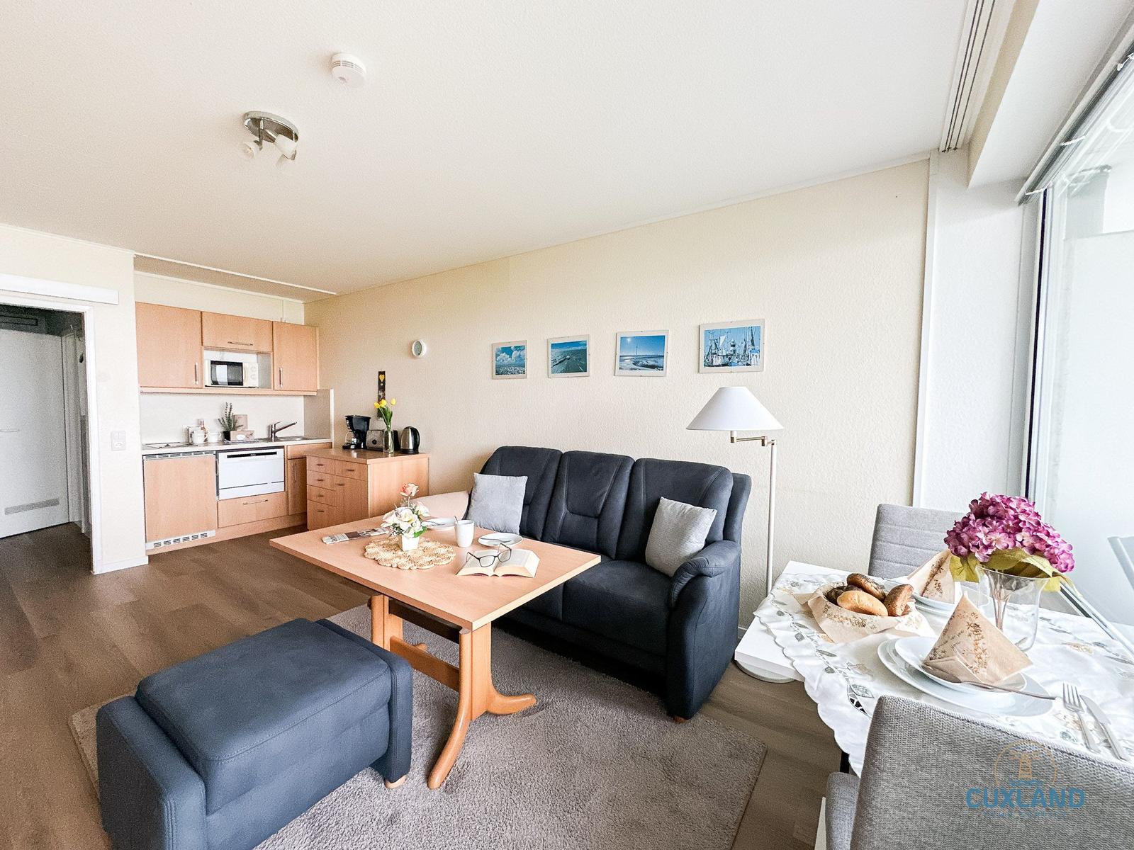 Wohnbereich mit Sofa, Esstisch und Küchenzeile. Große Fenster, Bilder an der Wand.