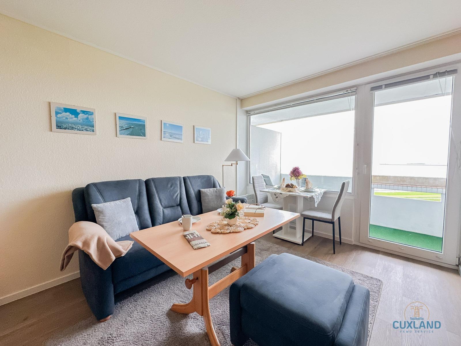Wohnbereich mit Sofa, Tisch und Balkon. An der Wand hängen Bilder.
