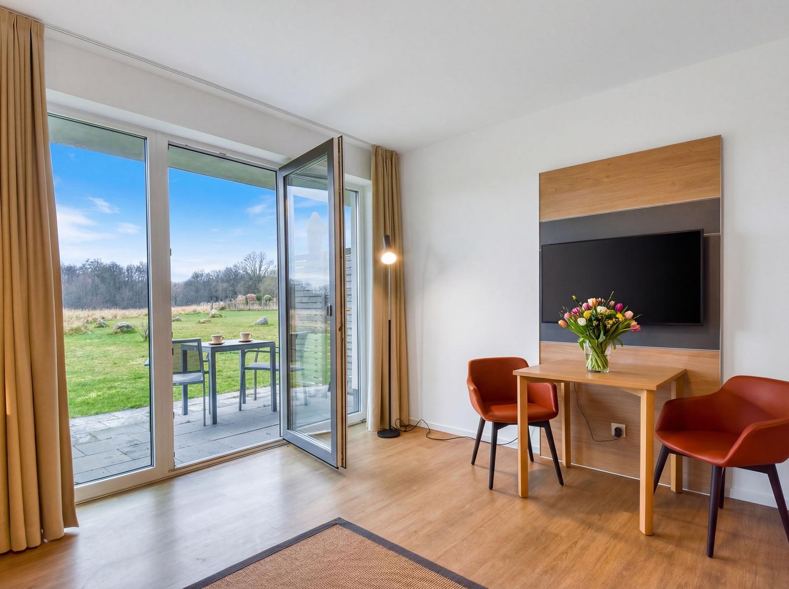 Ein Raum mit Tisch, zwei Stühlen und Fernseher. Durch die Glastüren sieht man einen Garten.