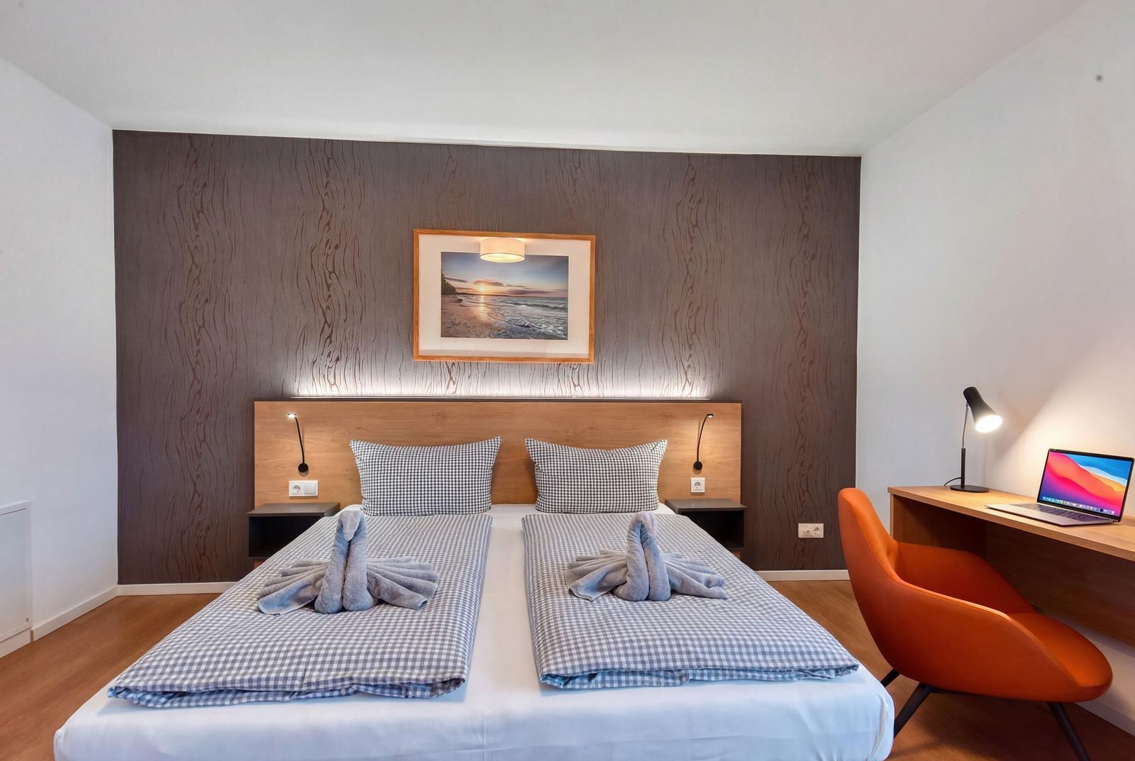 Doppelbett mit Kissen und Handtüchern, Schreibtisch mit Laptop und orange Stuhl.