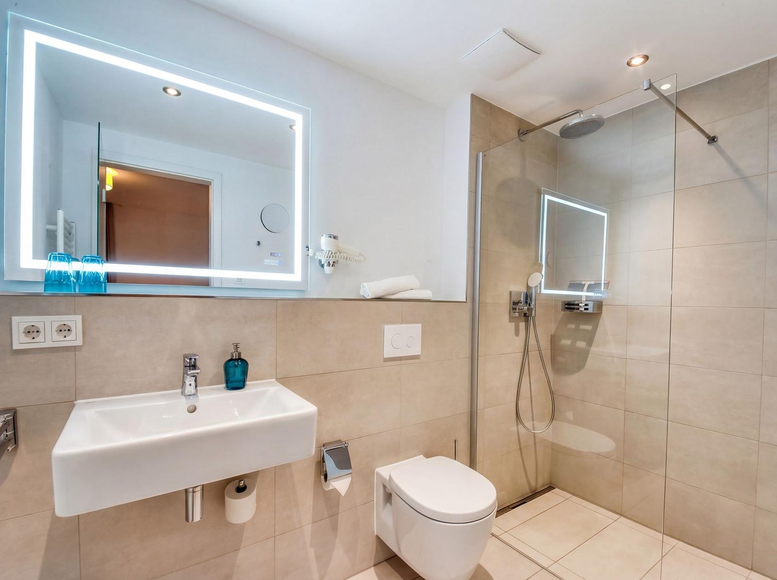 Badezimmer mit Dusche, Waschbecken und Toilette. Spiegel mit Beleuchtung und Handtuch.