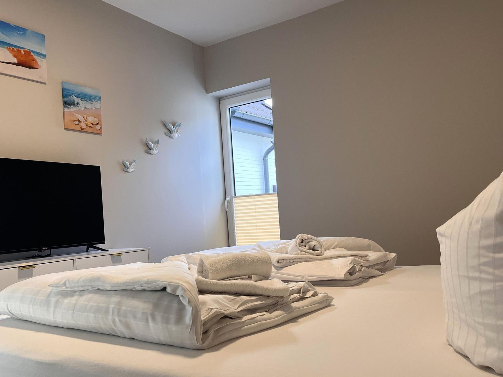 Ein Schlafzimmer mit Bett, Fernseher und Fenster. Wände sind hell, Bilder und Vogel-Dekoration sind sichtbar.