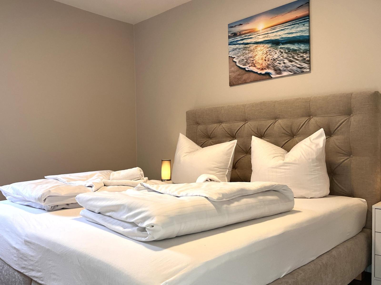 Ein Bett mit weißer Bettwäsche und einem braunen Kopfteil. An der Wand hängt ein Strandbild.