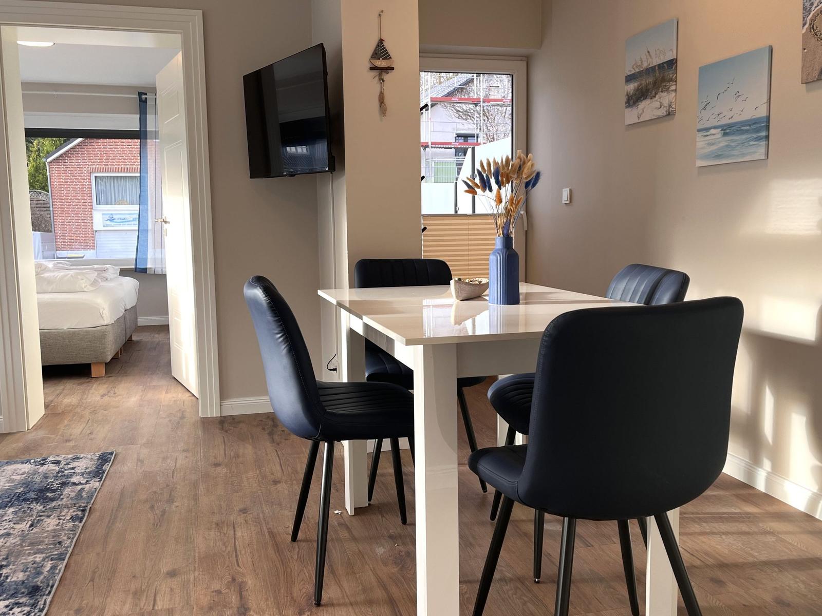 Essbereich mit Tisch, vier Stühlen und Fernseher. Im Hintergrund Schlafzimmer mit Bett.