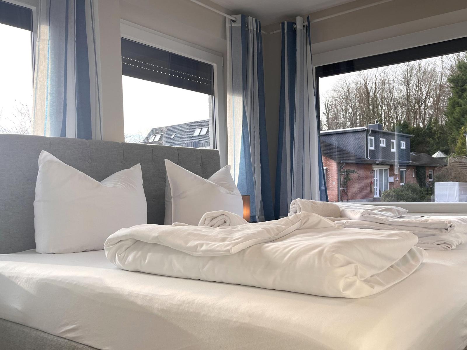 Das Bett mit weißen Laken und Kissen. Durch Fenster ist Außenansicht mit Häusern und Bäumen sichtbar.
