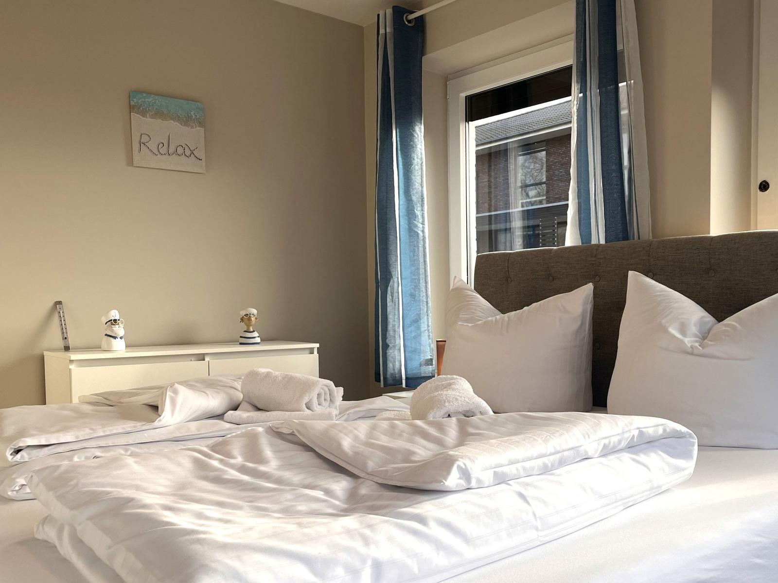 Ein Bett mit weißer Bettwäsche, Kissen und Handtüchern. An der Wand hängt ein Bild mit der Aufschrift Relax.