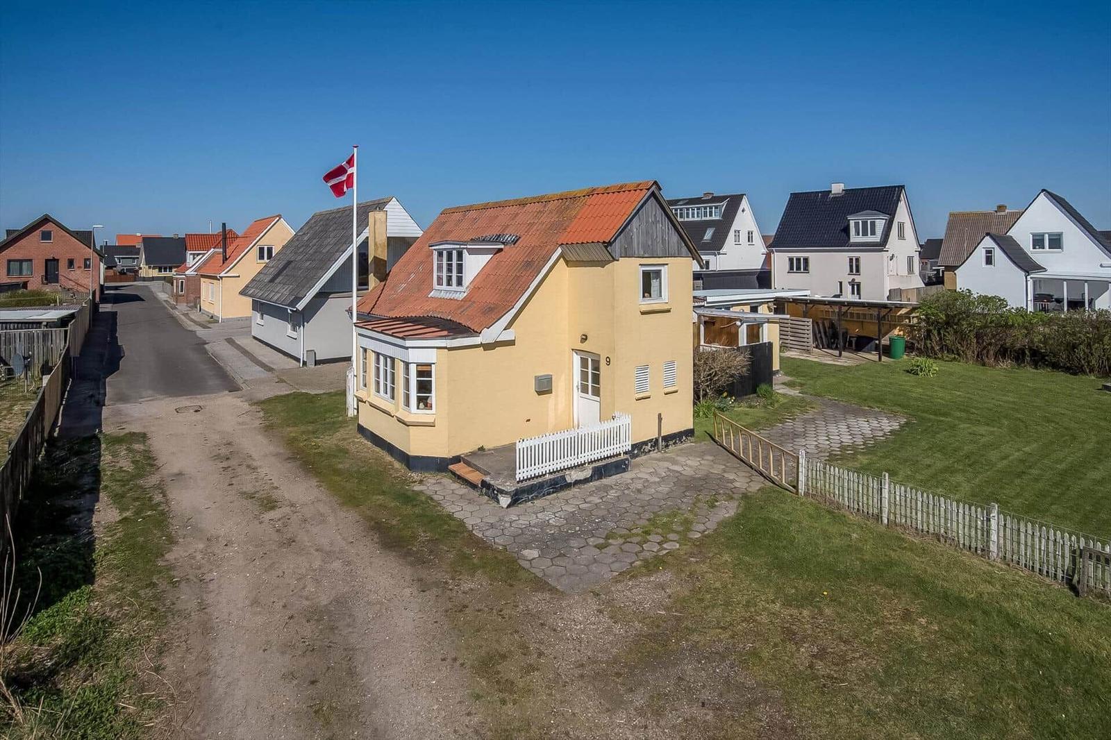 Yellow house with red roof, white fence, and garden. Other houses are nearby.