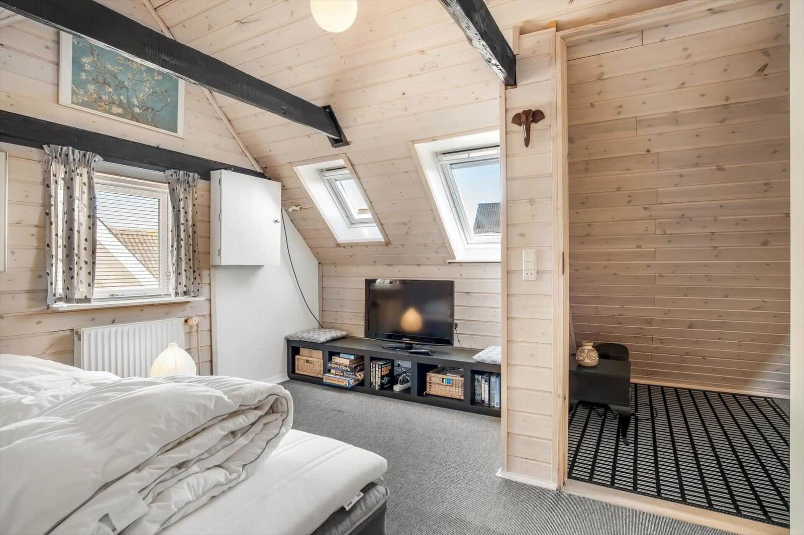 Bedroom with sloped ceiling, TV, and windows. Wooden walls and simple furnishings.