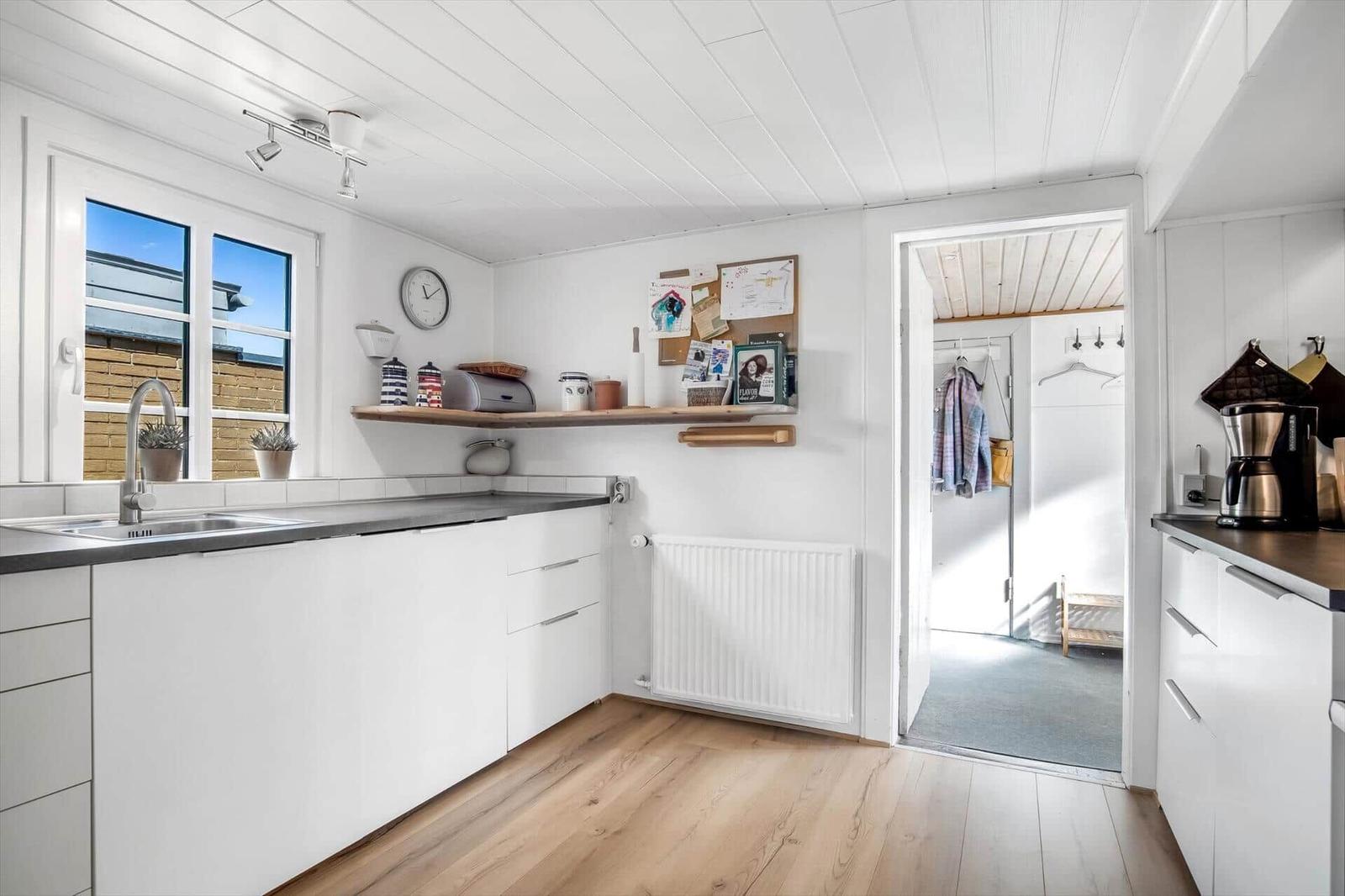 Kitchen with white cabinetry, wooden floor, and window. Passage to an adjacent room.