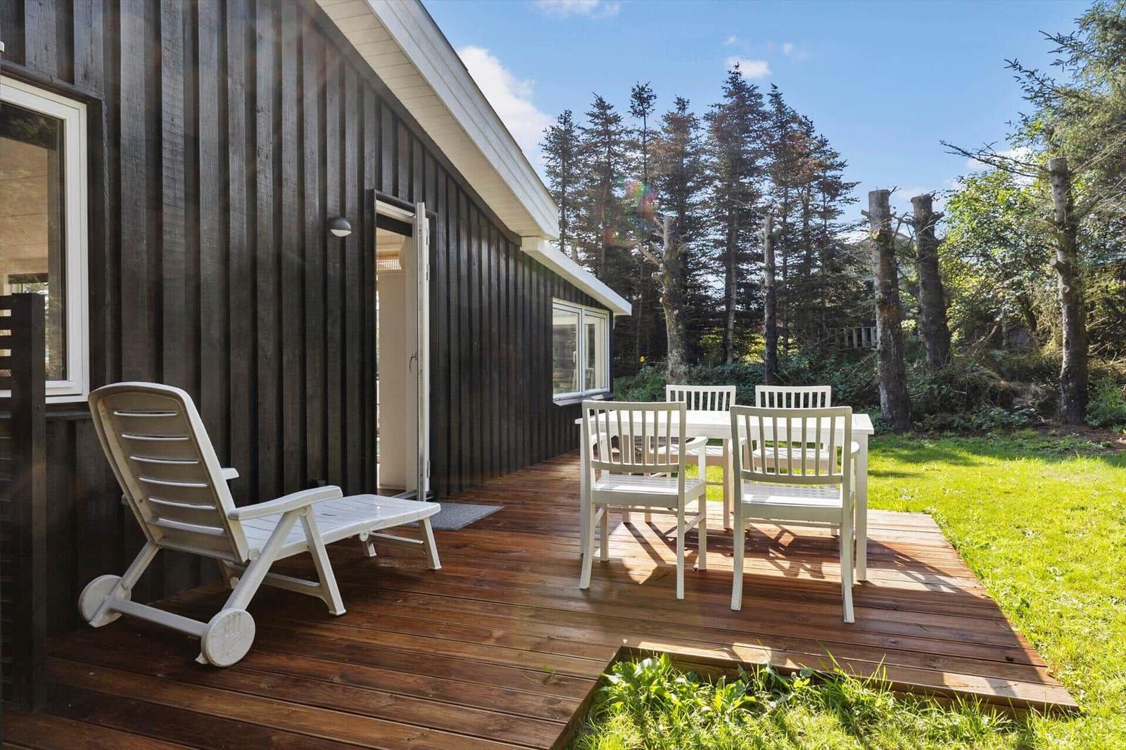 Deck with white table and chairs, lounge chair, and wooden flooring beside a house with dark siding.