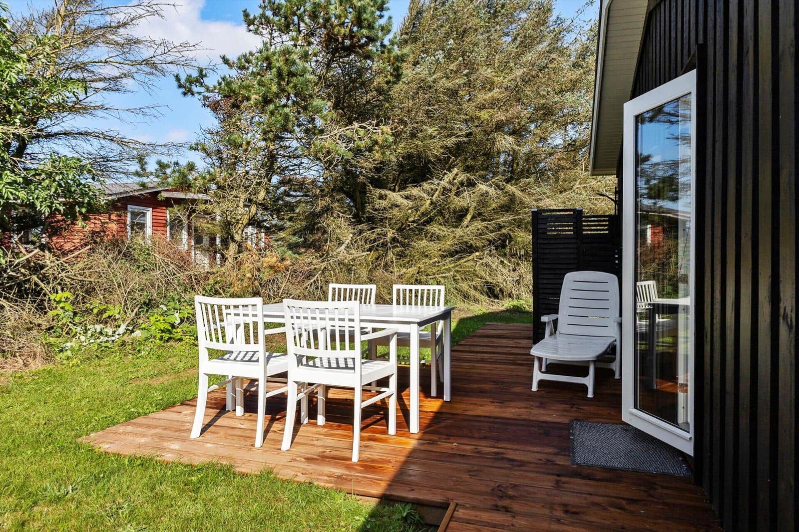 Deck with white table and chairs, wooden floor, adjacent garden and trees.