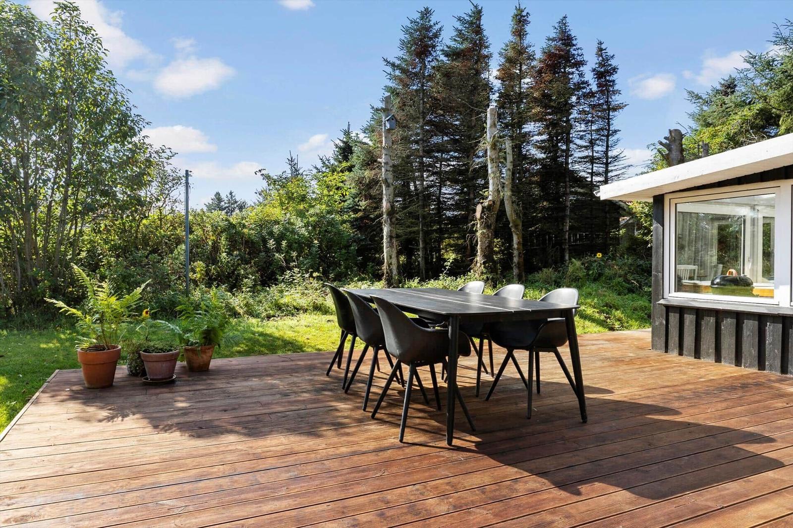 Deck with table and chairs, surrounded by greenery and trees. House with window on the right.