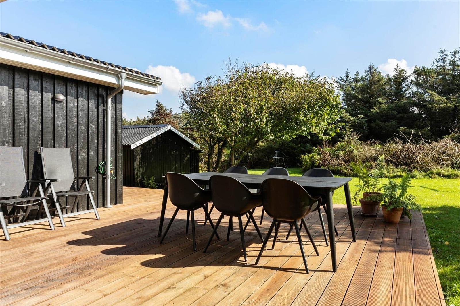 Deck with table and chairs, adjacent to house with garden and trees.