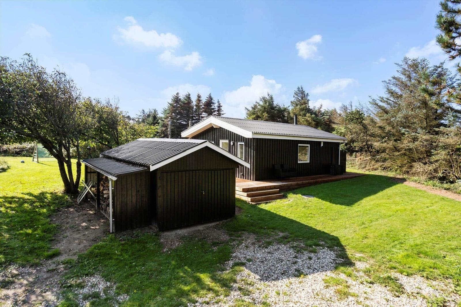 A dark-clad house with a terrace and extension. Surrounded by grass and trees under a blue sky.