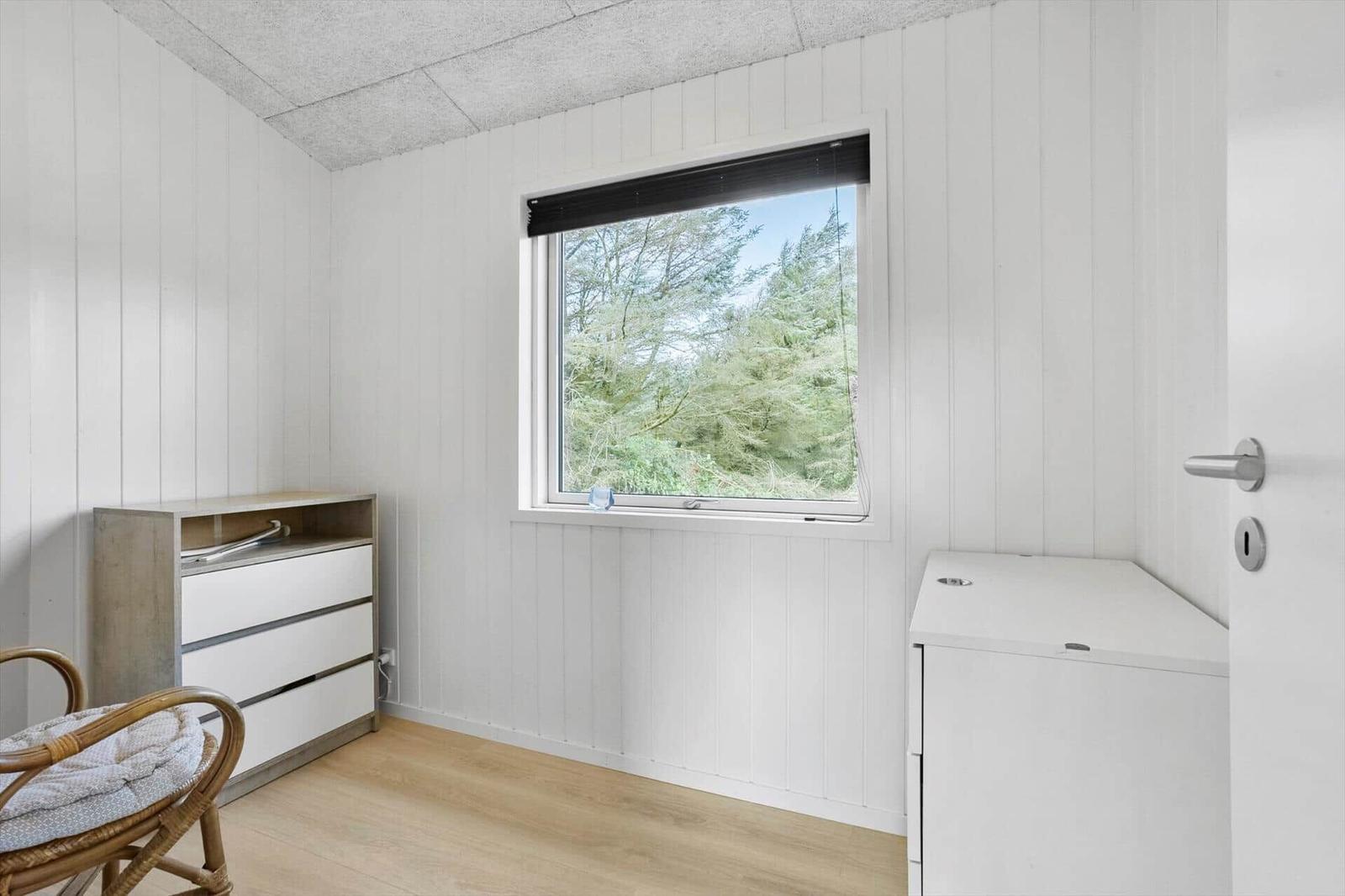 A room with white paneled walls, a window overlooking trees, and a dresser.