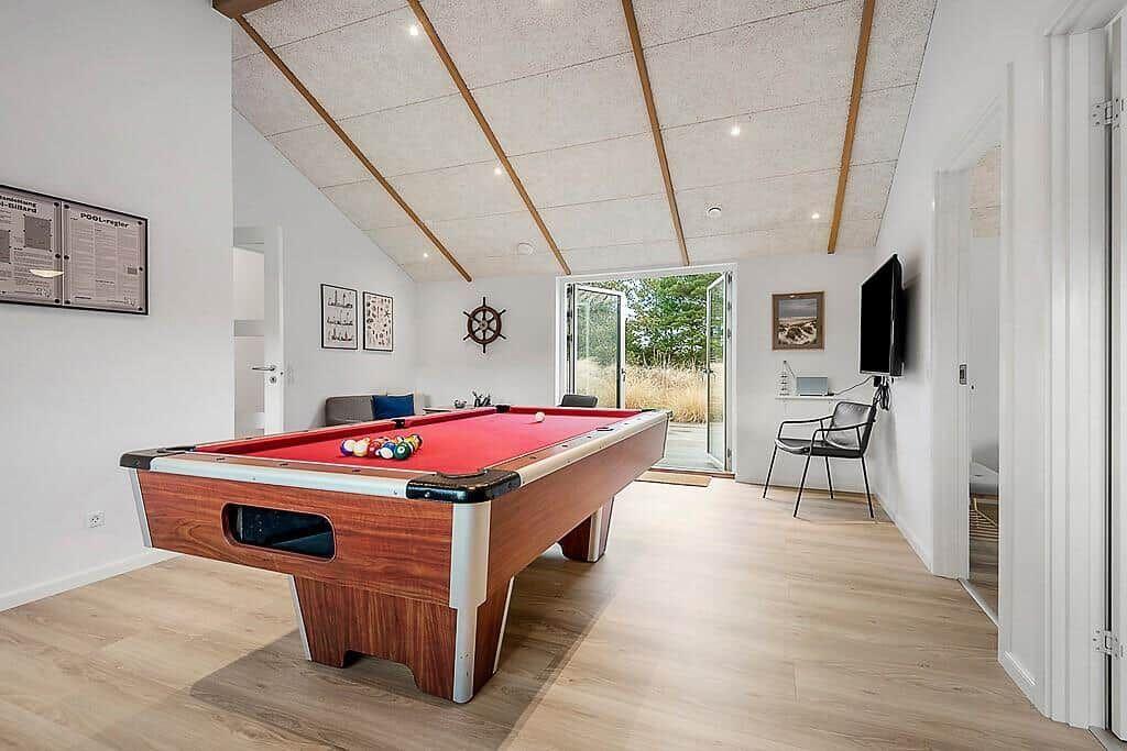 Billiard table with red felt, TV, and seating area in a bright room with wooden floor and sloped ceiling.