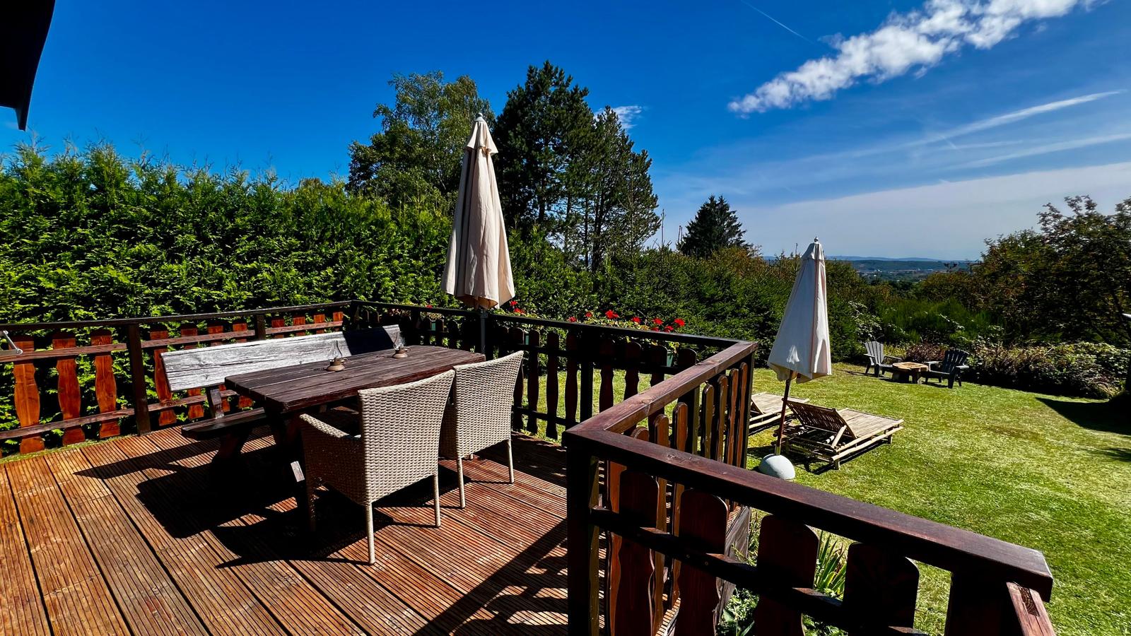 Terrasse mit Tisch, Stühlen und Sonnenschirmen. Blick auf grünen Garten und Wald.