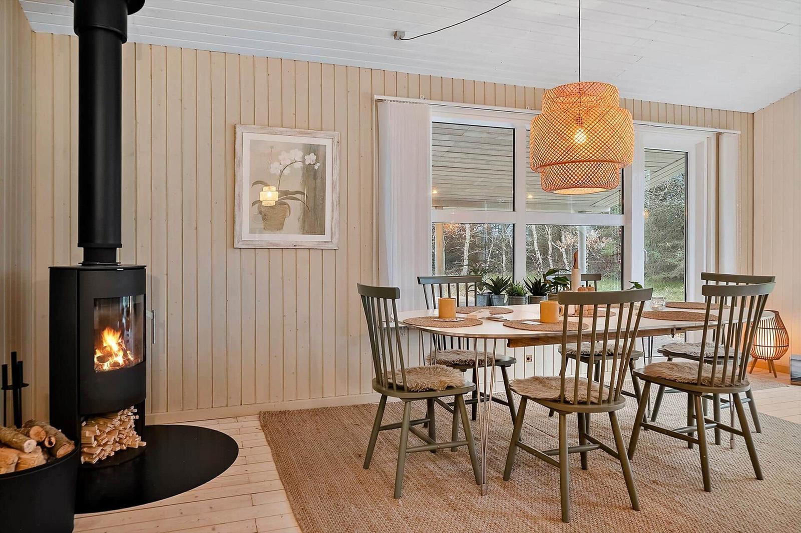 Esszimmer mit Holztisch, Stühlen und Kaminofen. Fenster zeigen Wald.