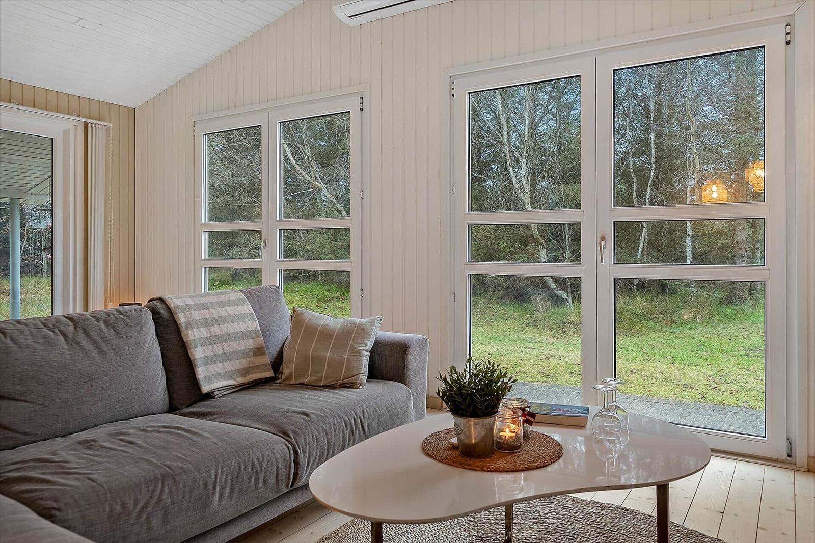Ein Wohnzimmer mit grauem Sofa, weißem Tisch und großen Fenstern zur Natur.