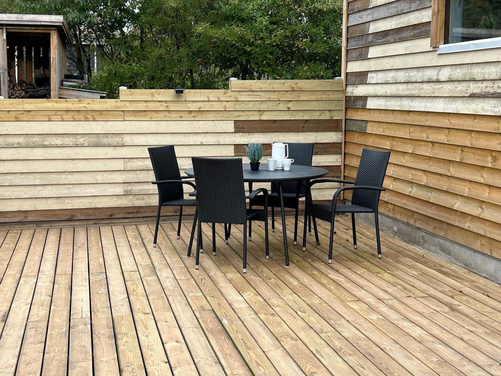 Deck with table and chairs, wooden wall, and garden in background.