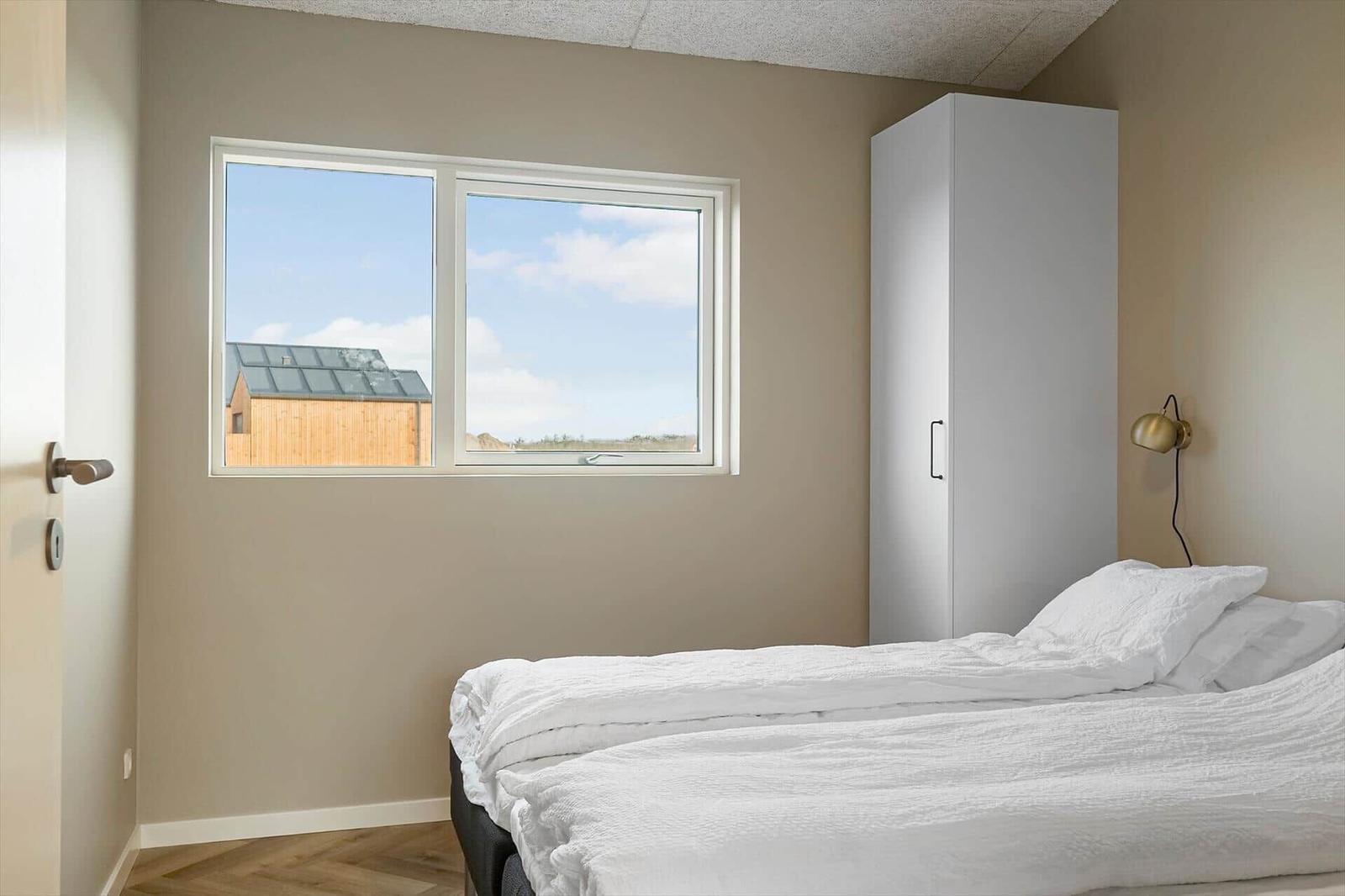 Schlafzimmer mit weißem Bett, Kleiderschrank und Fensterblick auf Holzhaus.