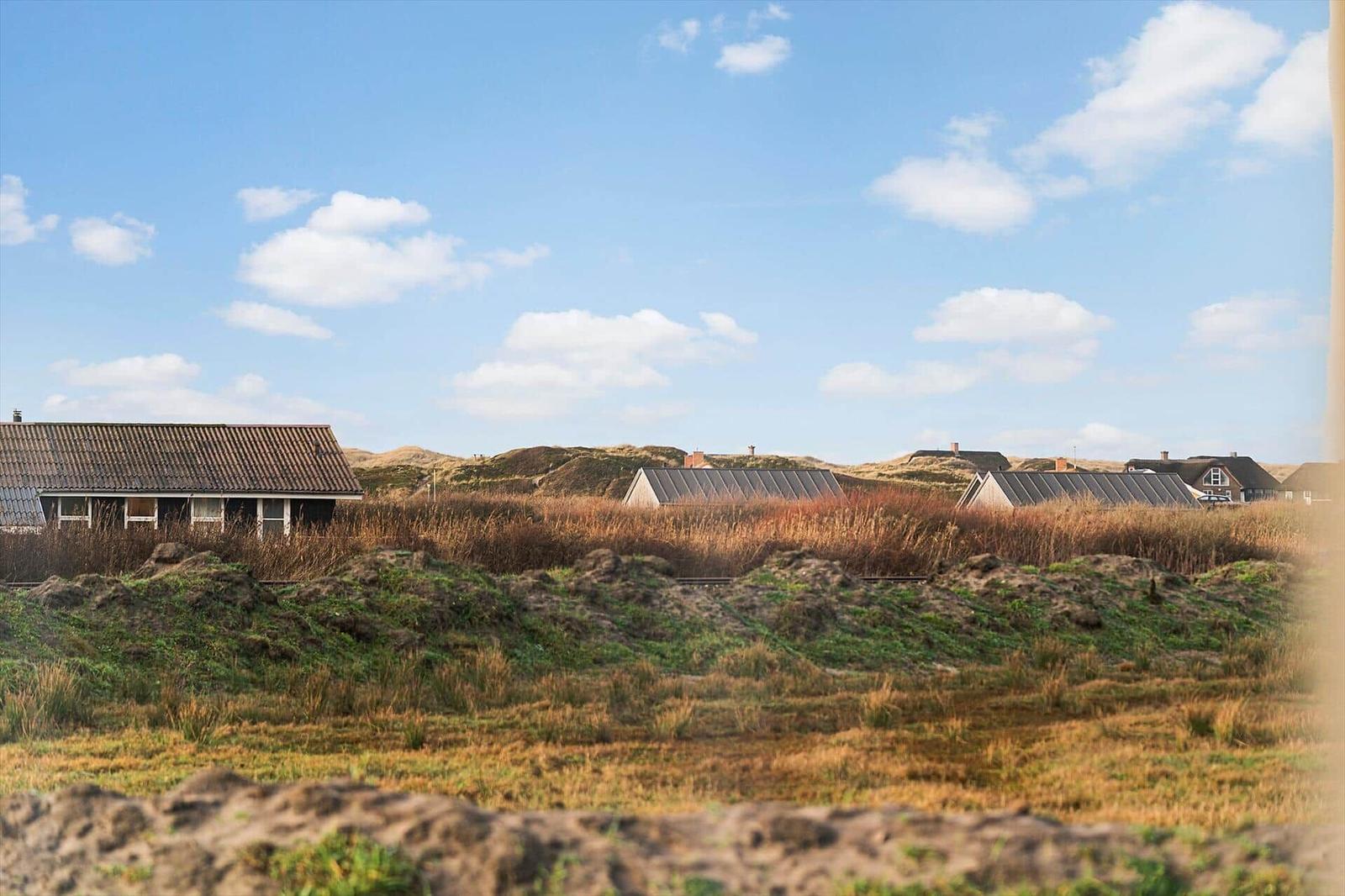 Häuser in der Ferne, umgeben von Dünen und Grasland unter blauem Himmel mit weißen Wolken.