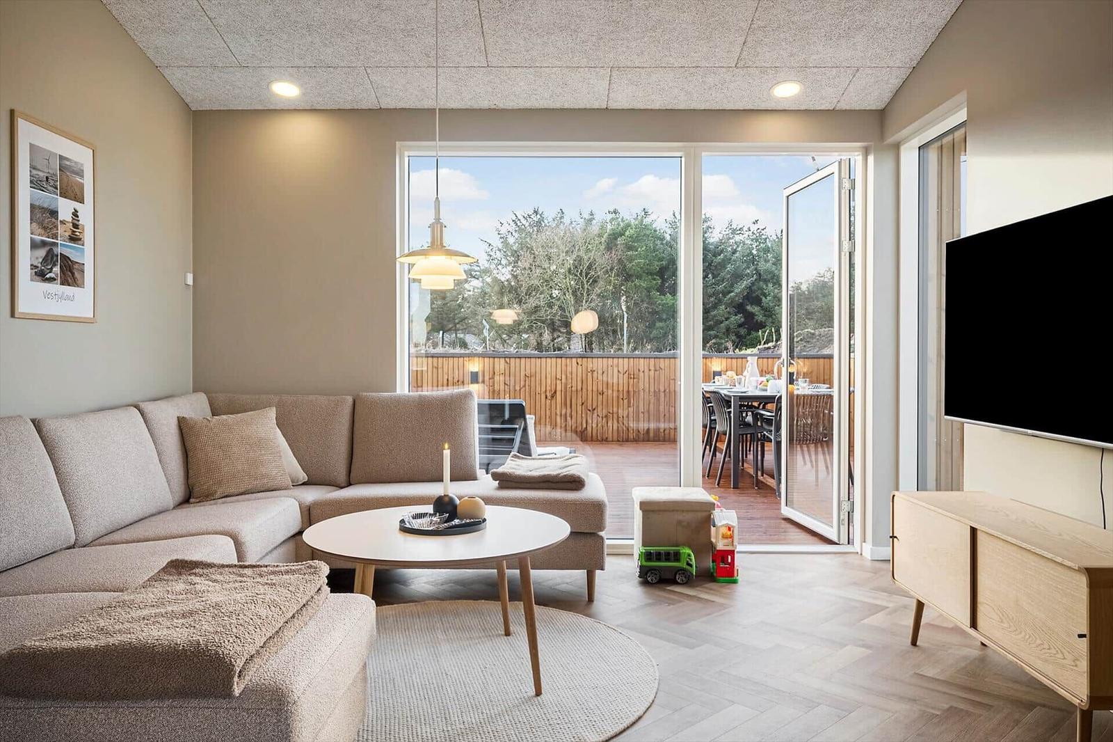 Wohnzimmer mit Sofa, Couchtisch und Fernseher. Durchgang zur Terrasse mit Tisch und Stühlen.