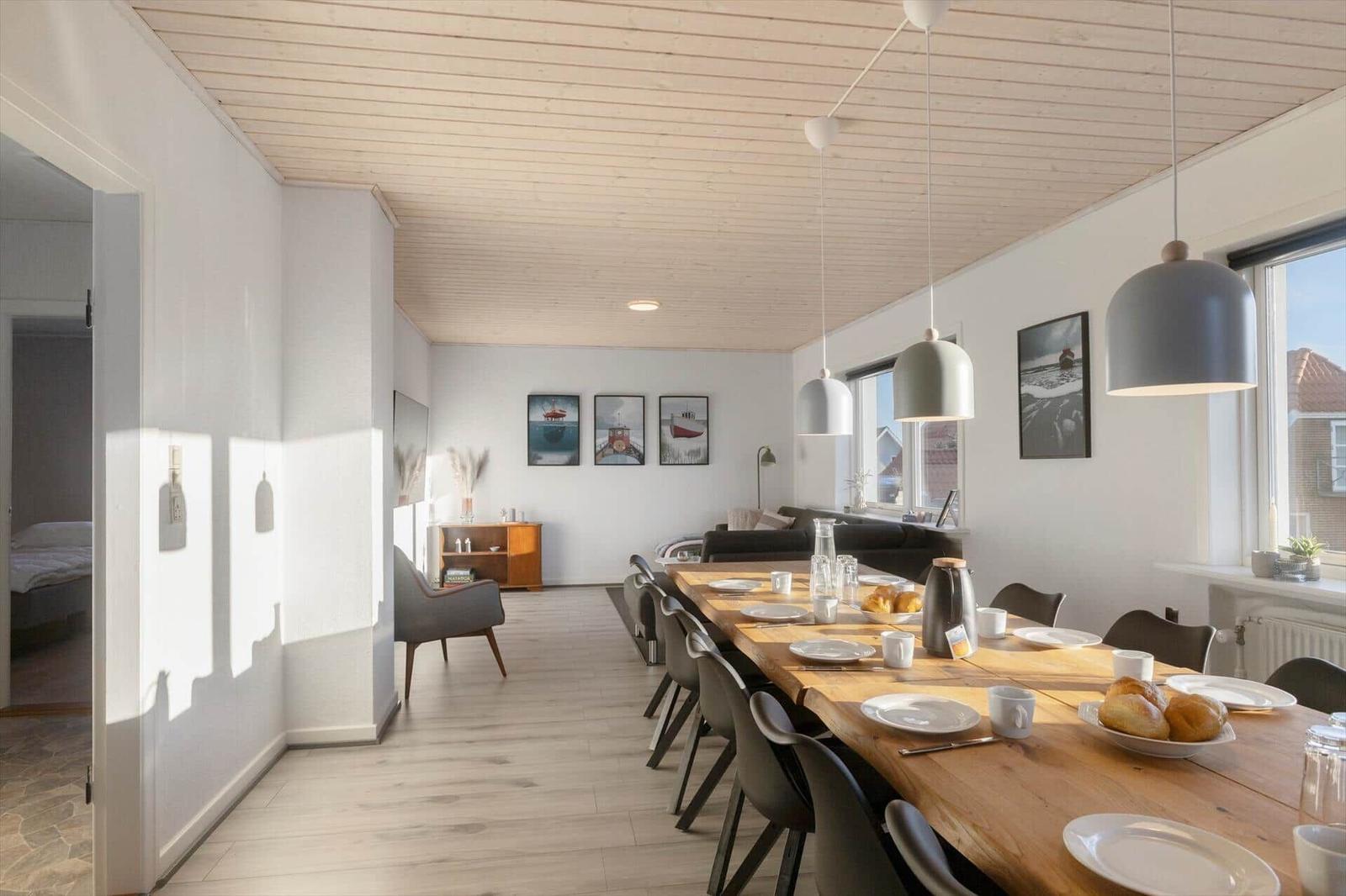 Dining room with long wooden table and chairs, living area in background, windows with view outside.