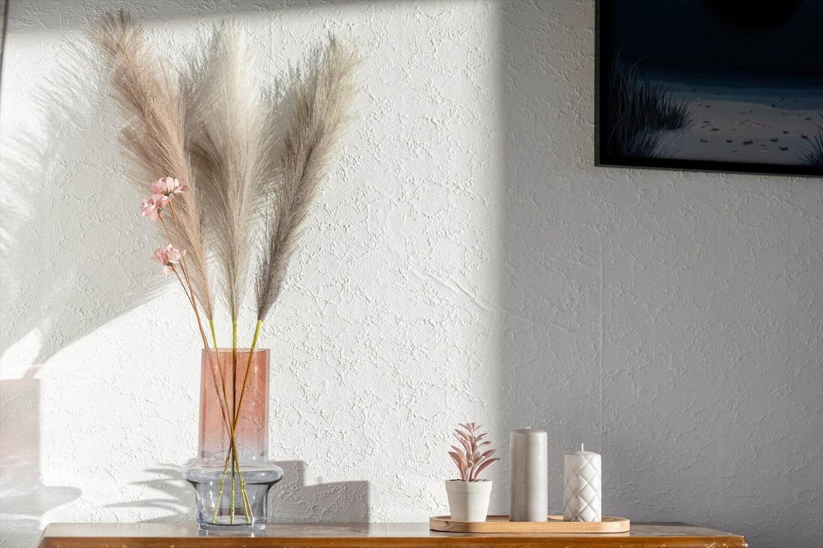 On a table are vases with pampas grass, a plant, and candles. A picture hangs on the wall.