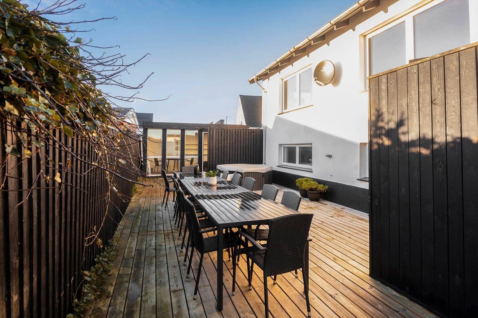 Terrace with long table and chairs, wooden floor, dark wood border, and garden house in background.
