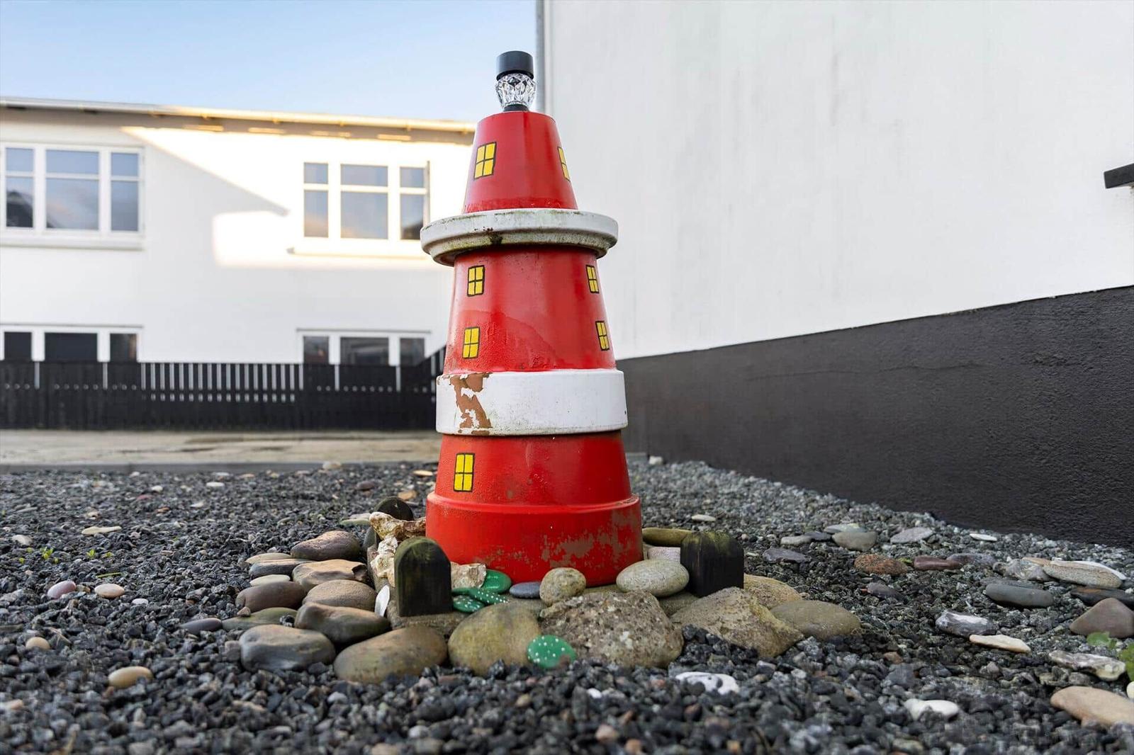 A red plastic lighthouse stands on stones in front of a house.