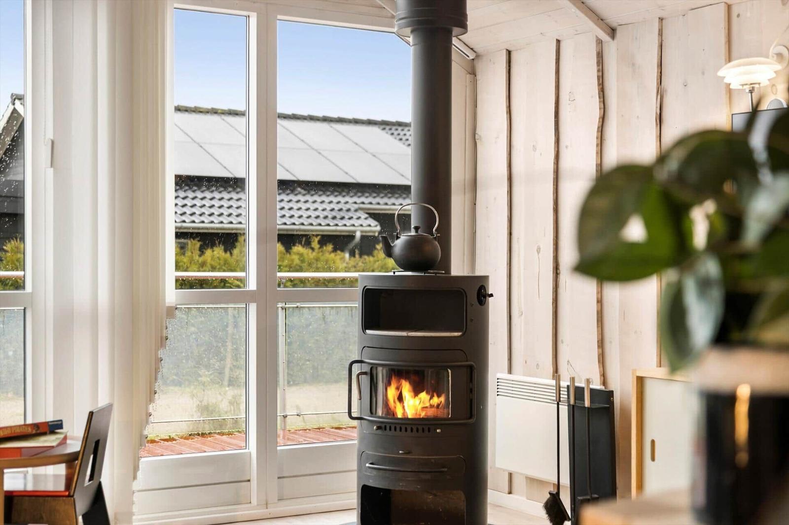 A wood stove with a kettle stands before large windows. Wooden walls and plants are visible.