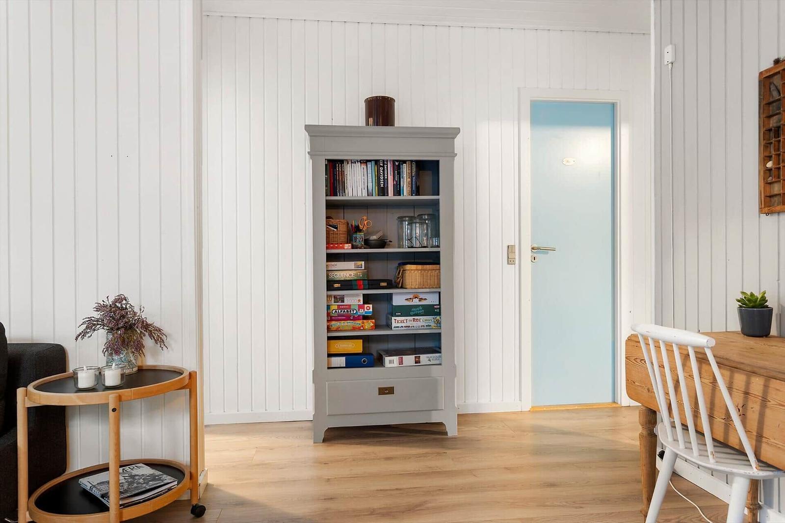 Ein Raum mit Bücherregal, Tisch, Stuhl und Tür. Wände sind weiß verkleidet.