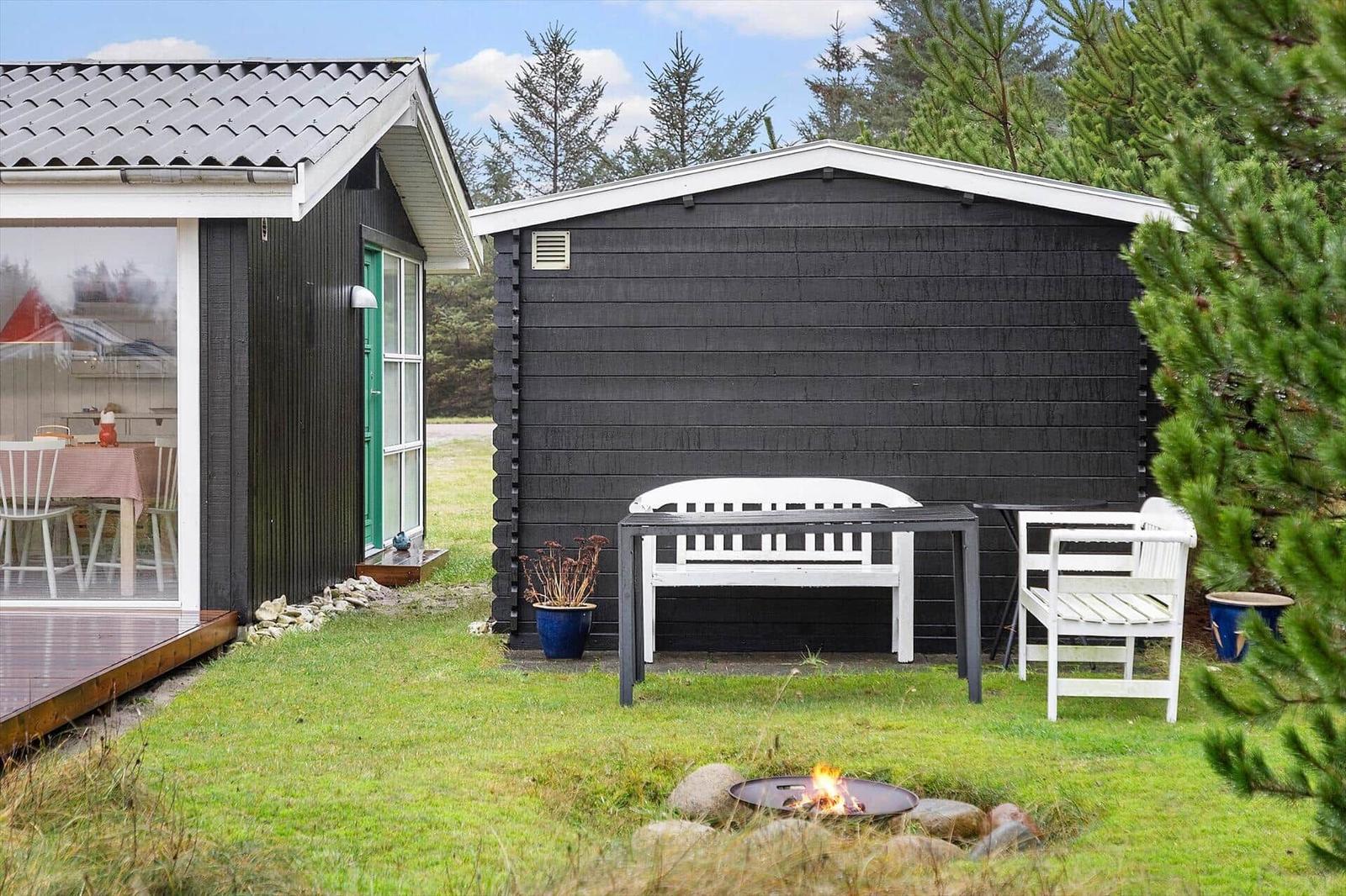 Schwarze Holzhütte mit weißem Gartenmöbel und brennendem Feuerkorb im Gras.