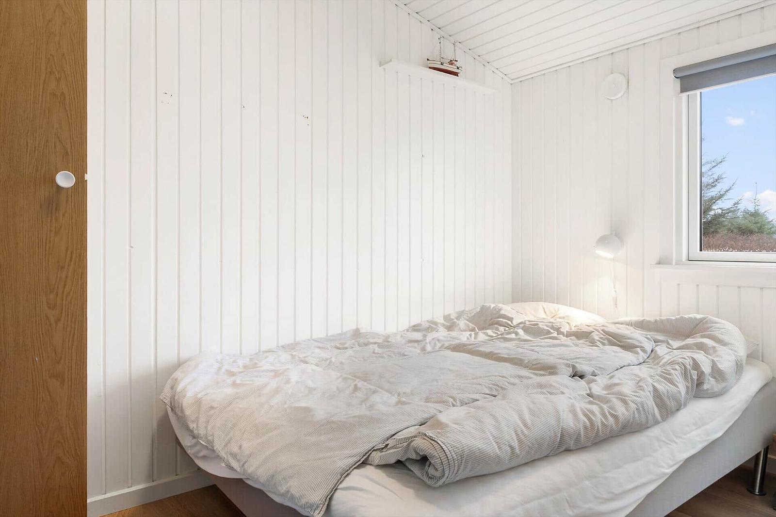 Schlafzimmer mit Bett, Fenster und Holzwand. An der Wand hängt eine Lampe.